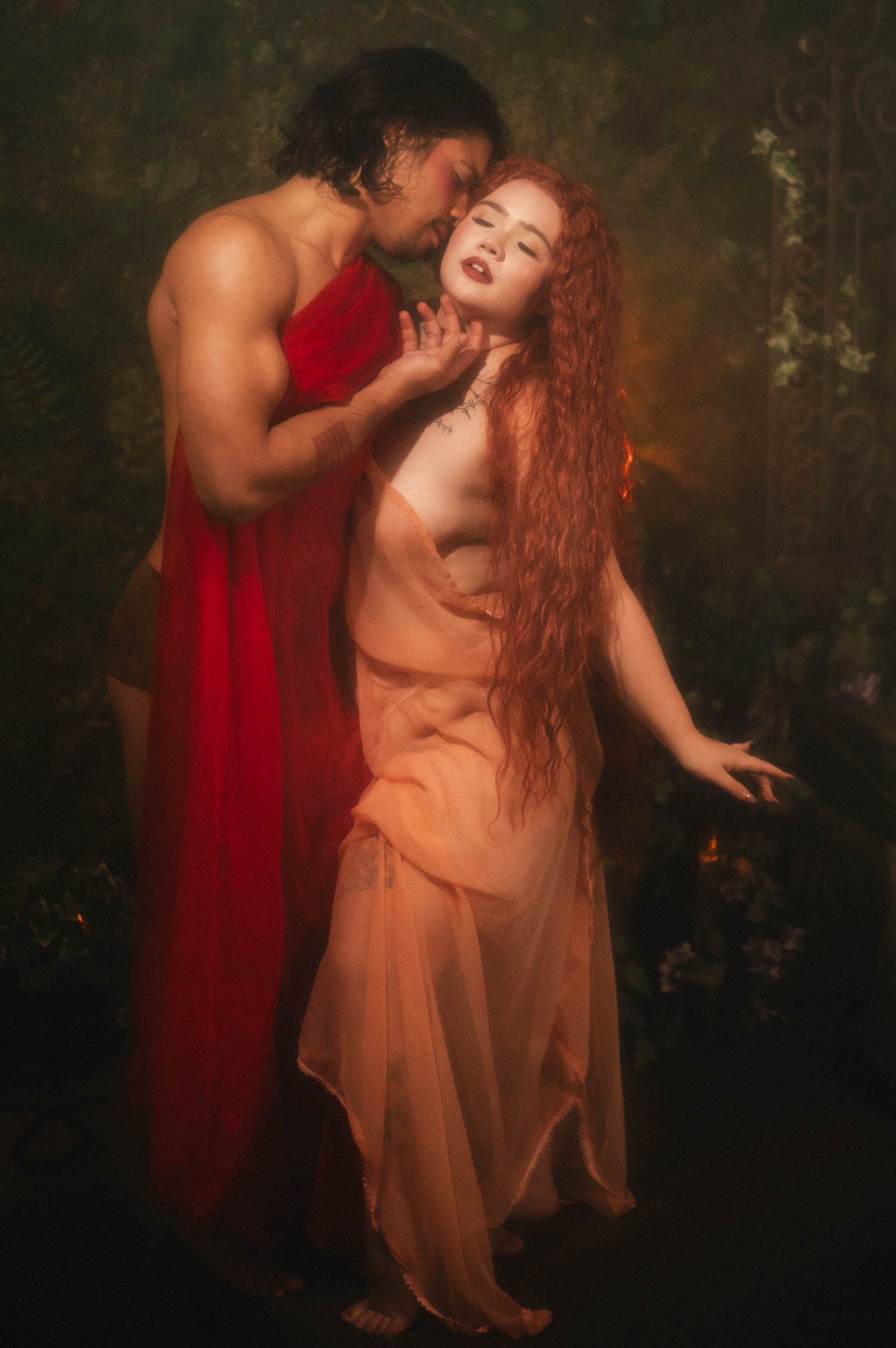 Statue inspired photoshoot moment shows the man kissing his partner's cheek as she reaches outward, her wet coral fabric catching warm firelight. Royal Lune Photo