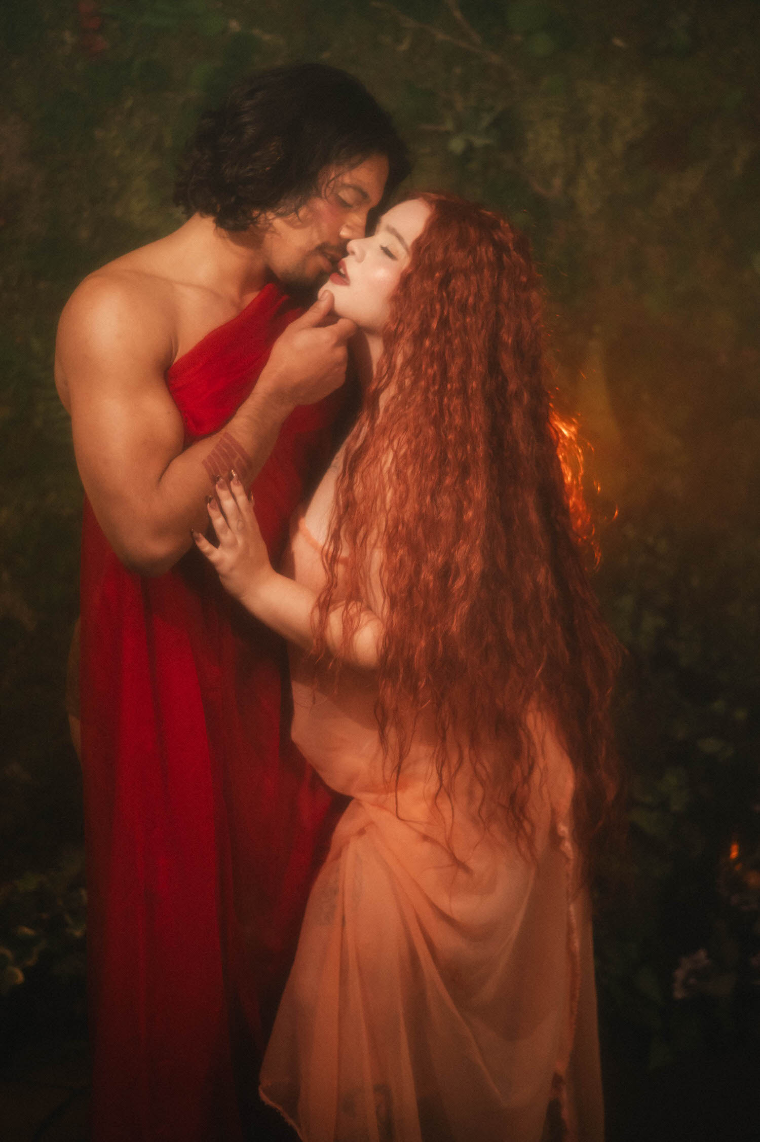 A Renaissance boudoir photoshoot kiss captured in profile, the woman's head tilted back as her partner leans in beneath dark hanging greenery. Royal Lune Photo