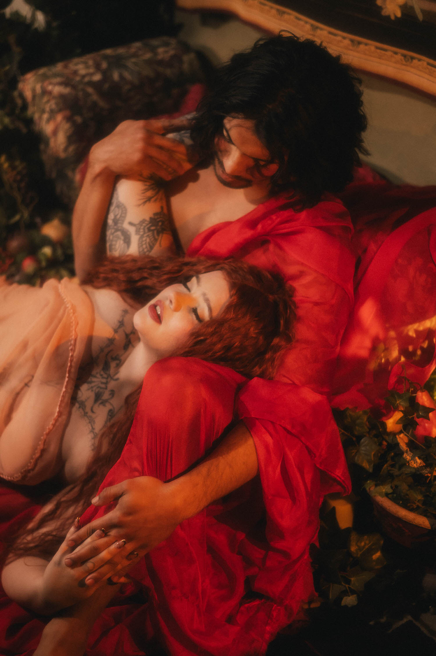 A statue inspired photoshoot pose shows a couple tangled together on a floral chaise, wet red silk pooling around them as the woman's tattooed leg wraps close. Royal Lune Photo