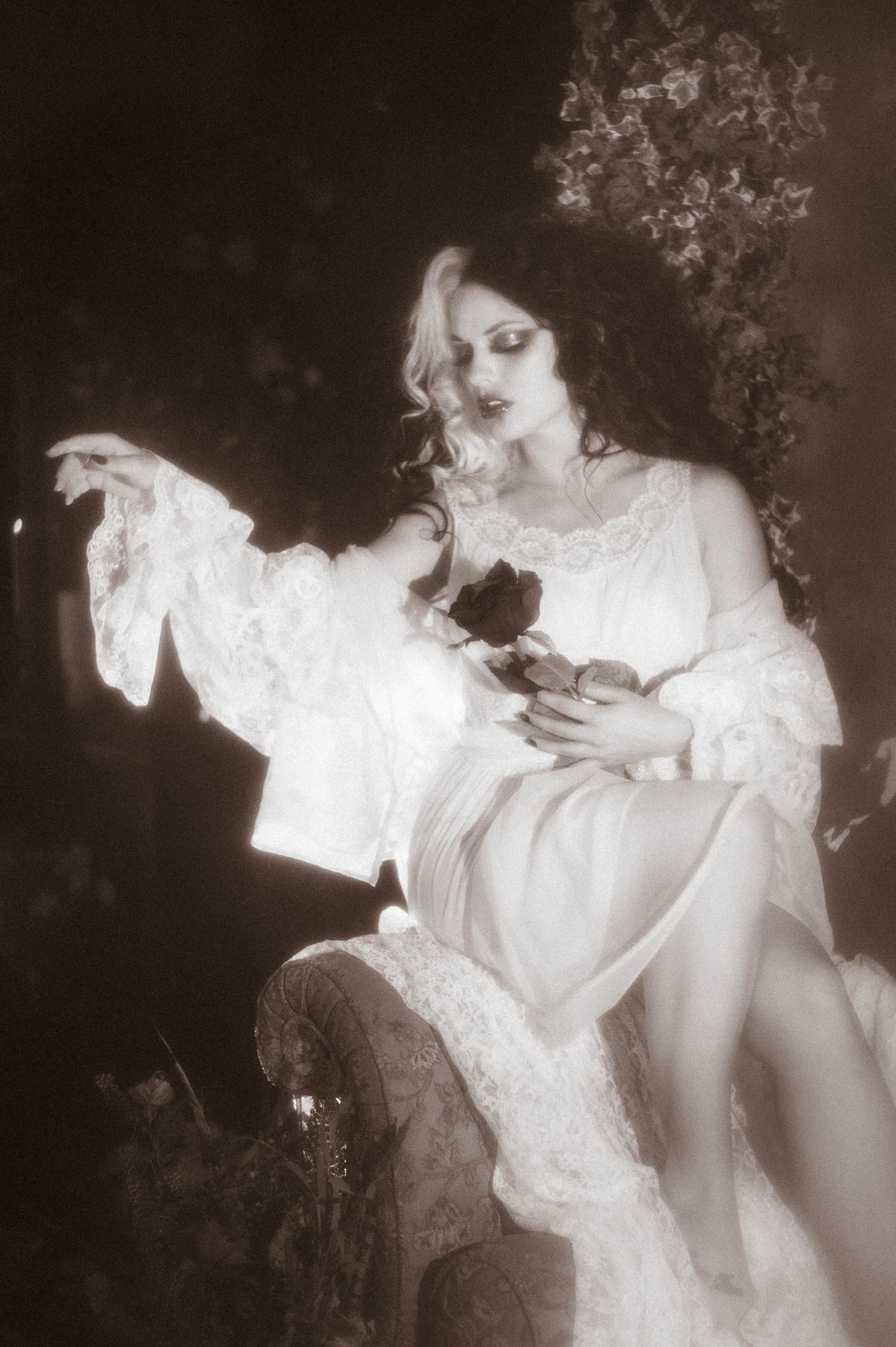 A model in a vintage sheer ivory lace dress sits holding a black rose while reaching one arm outward in this Tortured Poets Department photoshoot by Royal Lune Photo.