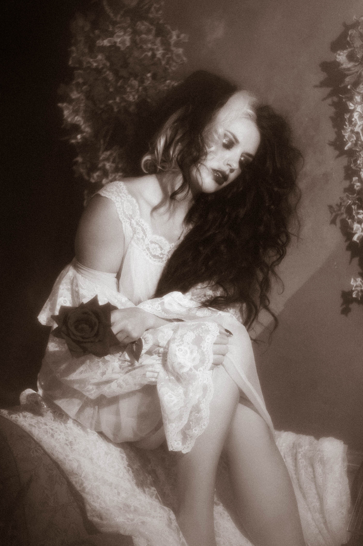 A black and white portrait shows a model seated in an ivory lace dress holding a dark rose against her knee in this gothic boudoir photography session by Royal Lune Photo.