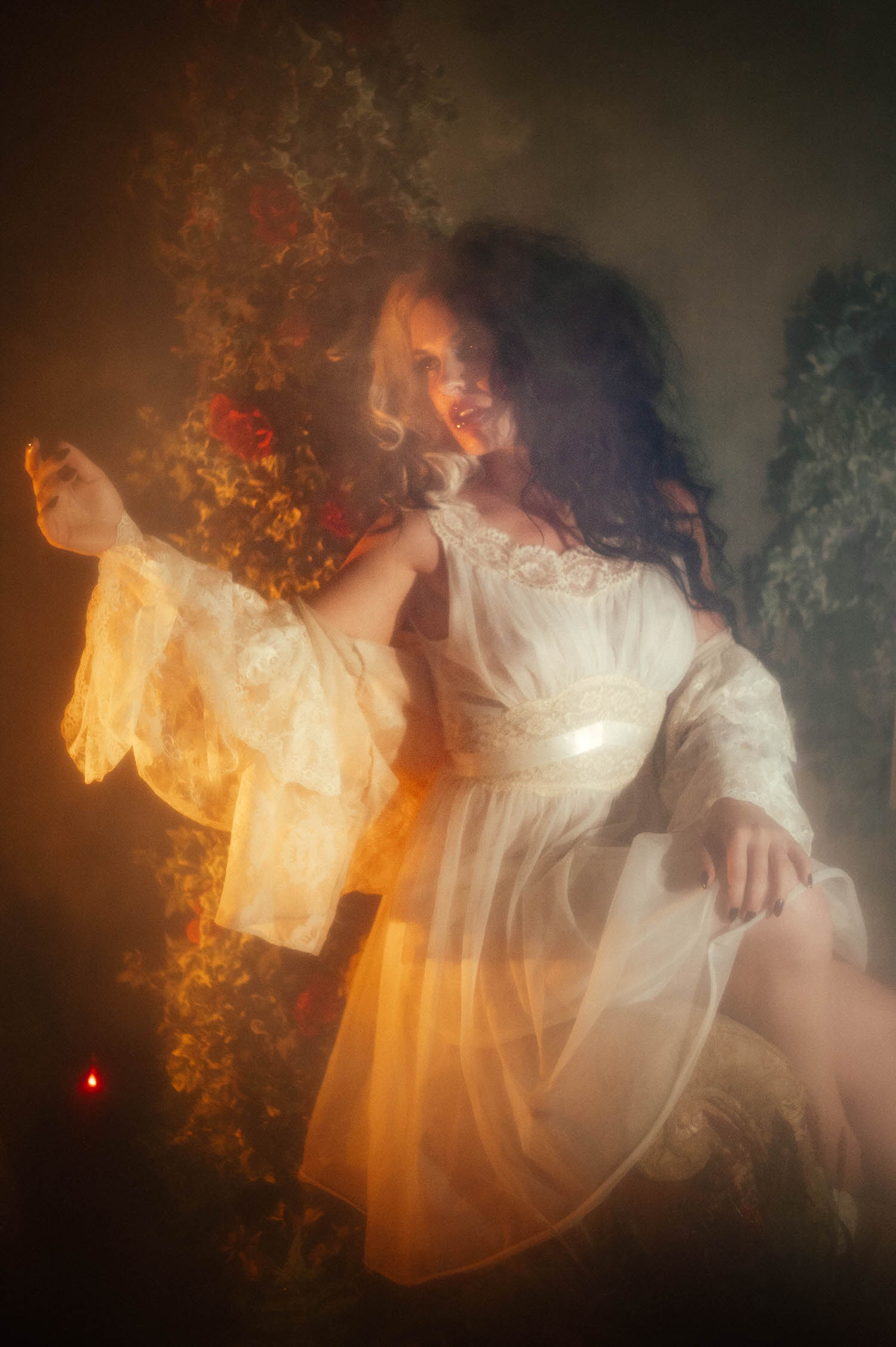 A model in a sheer ivory lace dress sits with one arm outstretched toward flickering candlelight and red roses in this dark academia photoshoot by Royal Lune Photo.