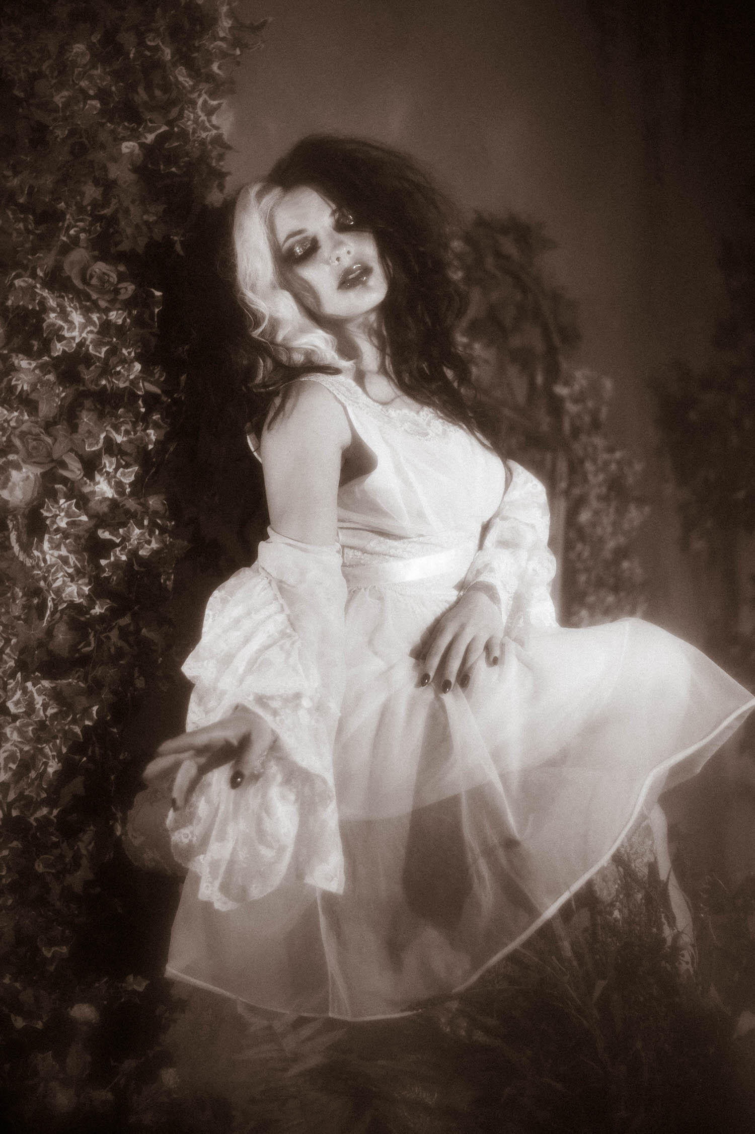 A black and white portrait shows a model in a sheer ivory lace dress seated with fabric caught mid movement beside climbing ivy and roses in this gothic boudoir photography portrait by Royal Lune Photo.