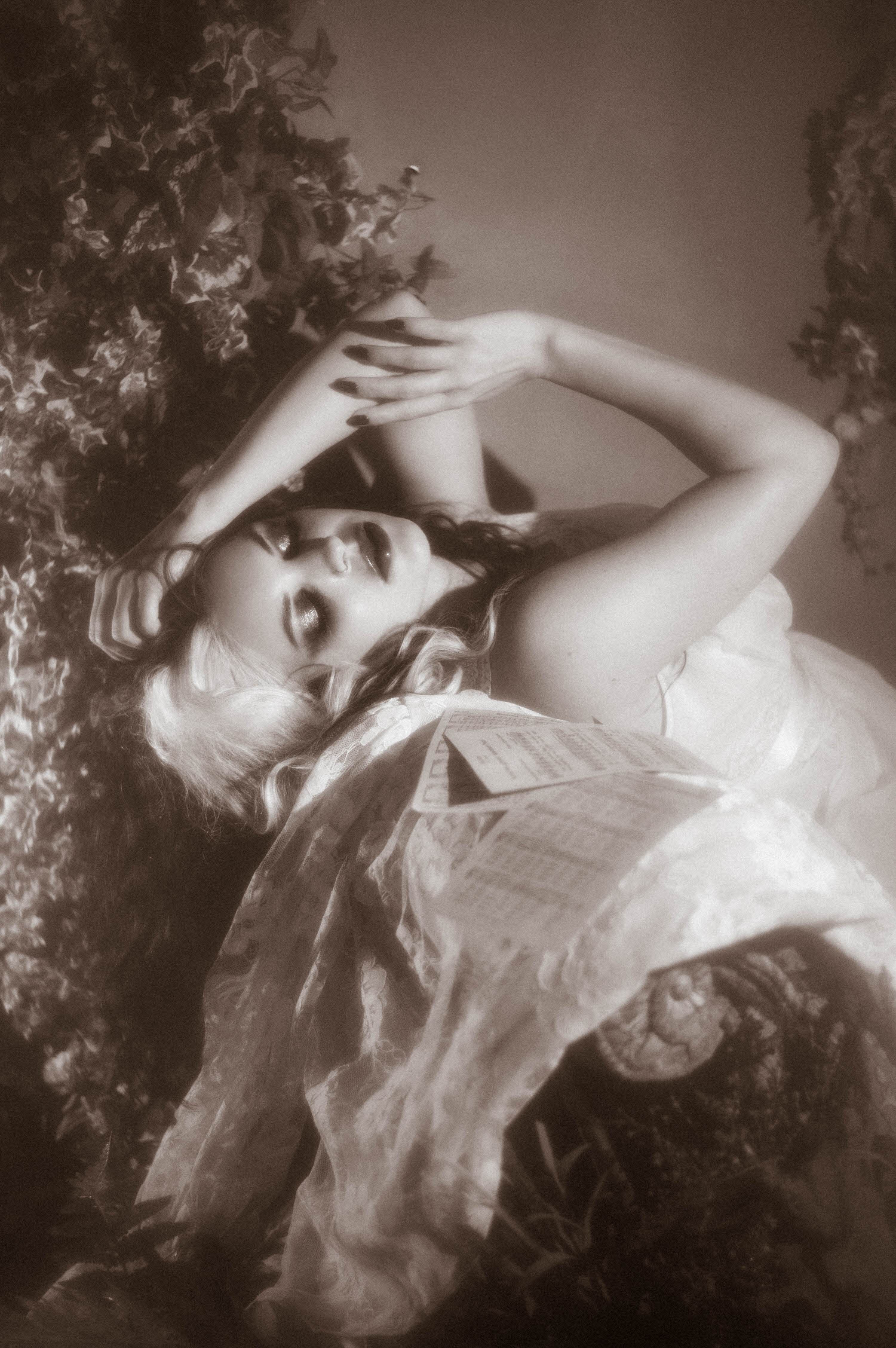A black and white portrait shows a model reclining with a page of sheet music resting against her, framed by ivy leaves in this fine art boudoir Dallas session by Royal Lune Photo.