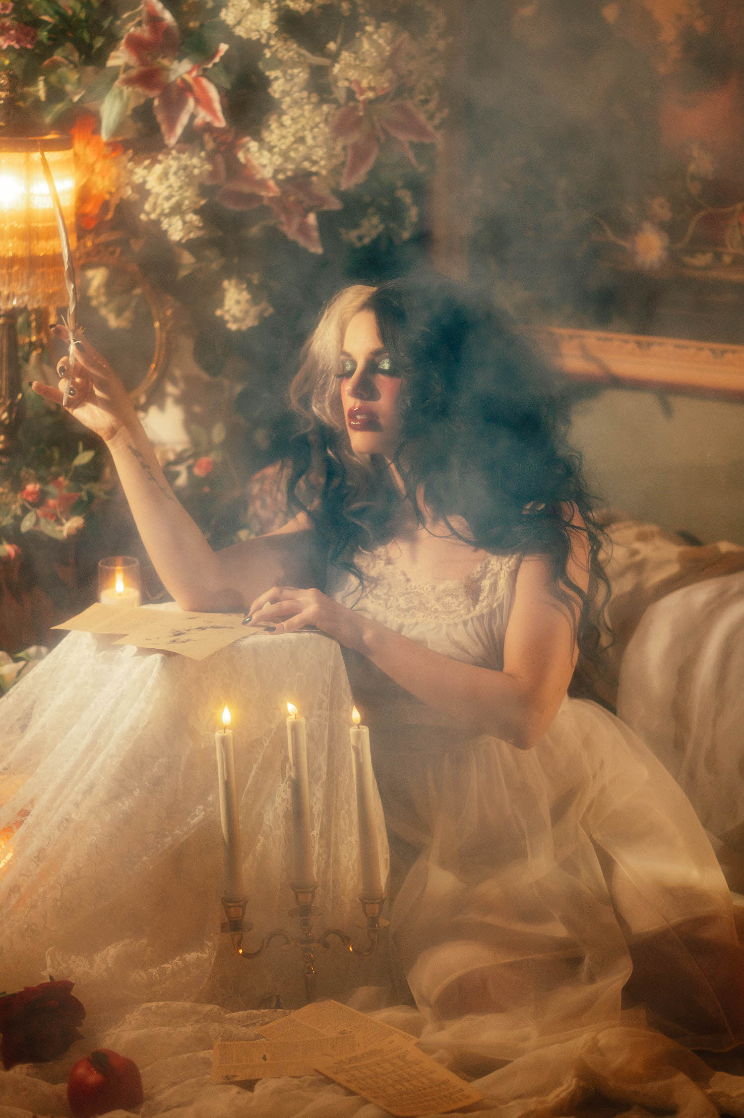 A model in a vintage ivory lace nightgown sits at a lace covered table raising a quill pen, surrounded by candlelight and soft fog in this Tortured Poets Department photoshoot by Royal Lune Photo.