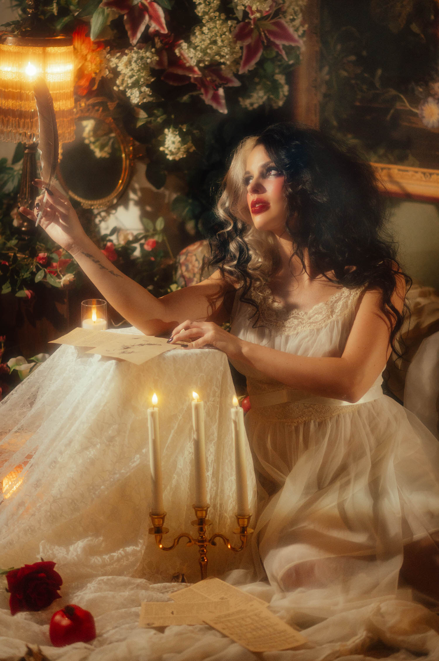 A model in an ivory lace nightgown sits at a candlelit table holding a quill pen above handwritten letters, framed by lush florals in this moody poetry photoshoot by Royal Lune Photo.