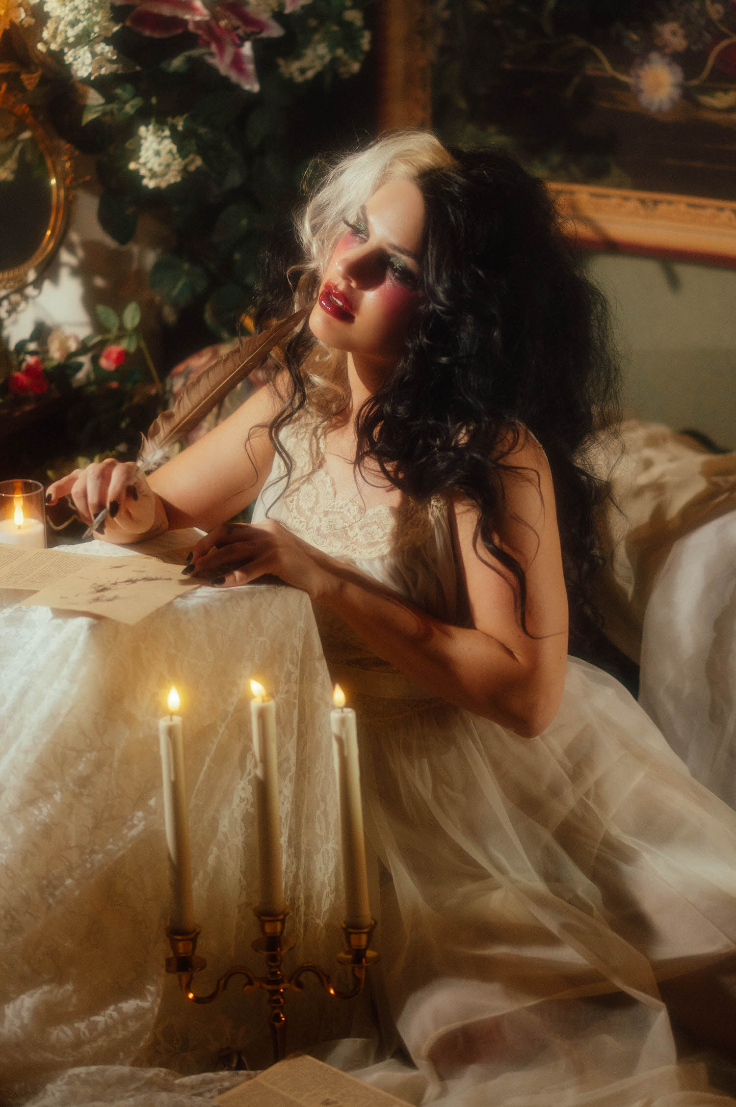 A model in an ivory lace nightgown rests a quill pen near her chin while seated at a candlelit writing table in this heartbreak photoshoot idea by Royal Lune Photo.