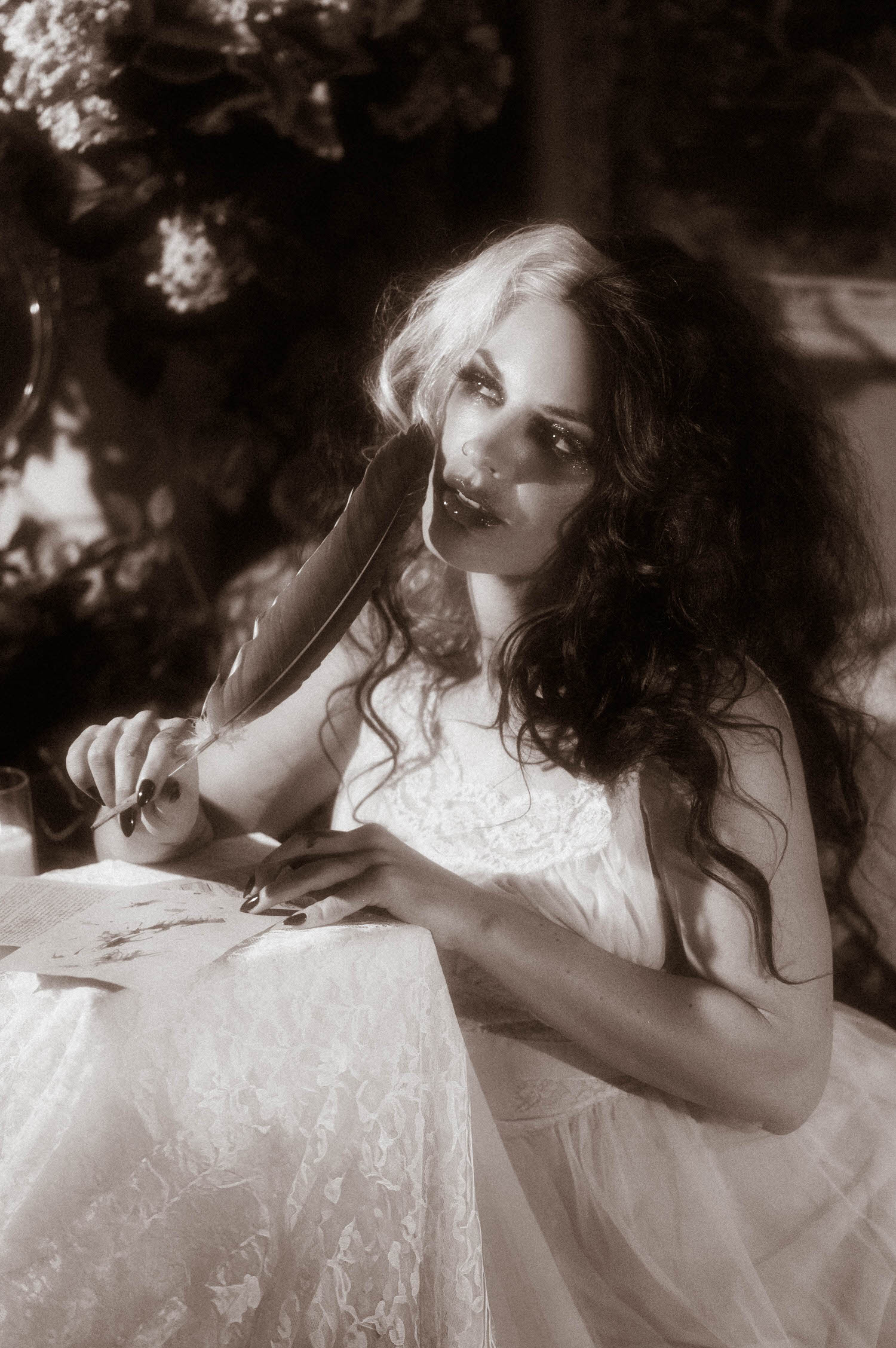 A black and white portrait shows a model in an ivory lace nightgown resting a quill pen against her cheek at a candlelit table in this gothic boudoir photography session by Royal Lune Photo.