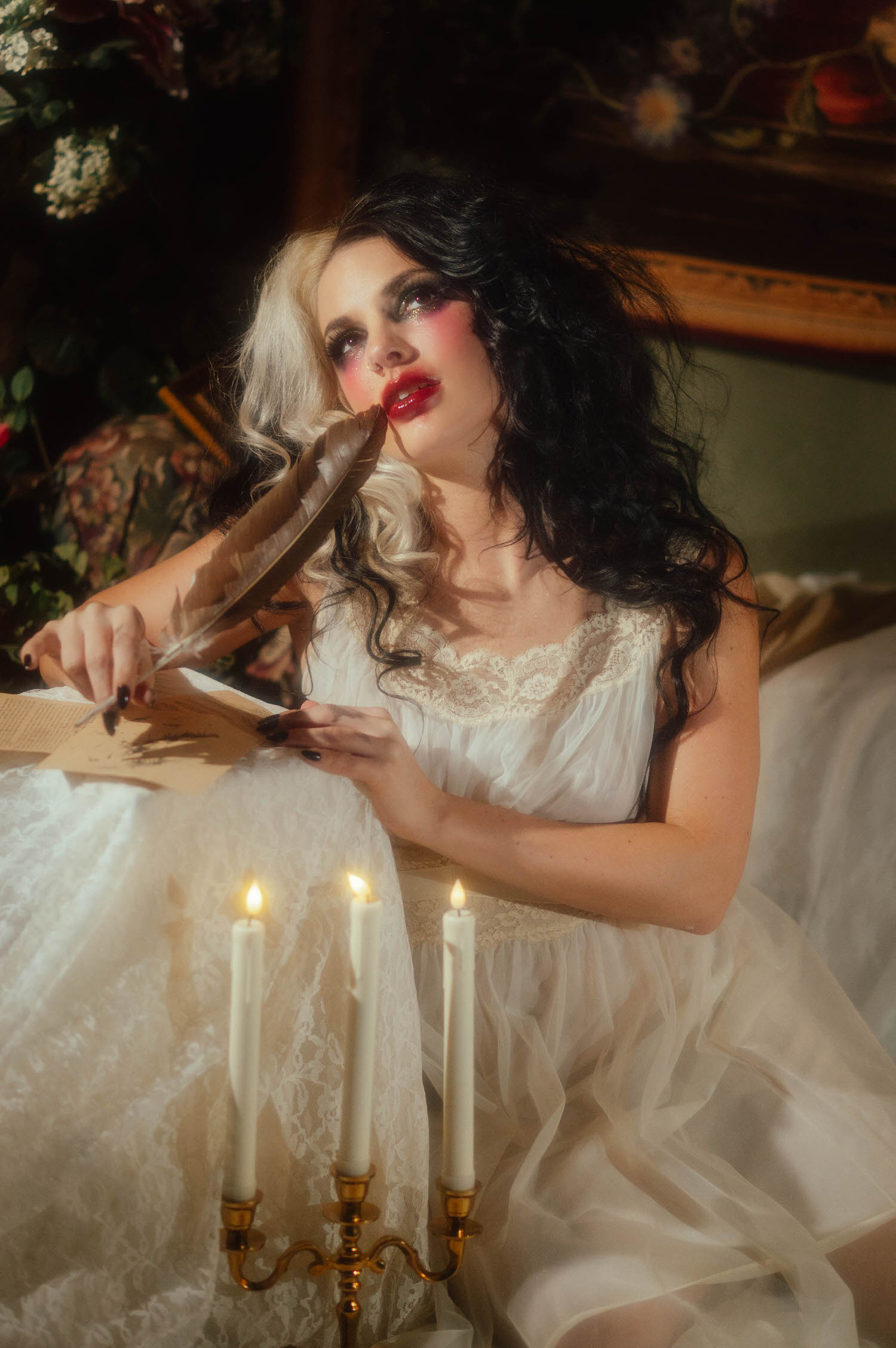 A model in an ivory lace nightgown holds a quill pen near her lips while seated at a candlelit table with handwritten letters in this Tortured Poets Department photoshoot by Royal Lune Photo.