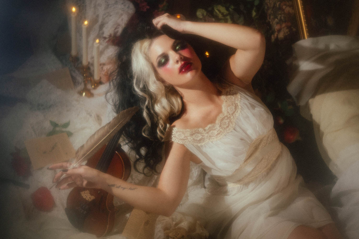 A model in an ivory lace nightgown reclines with one arm raised, holding a quill pen near a violin surrounded by candlelight in this gothic boudoir photography portrait by Royal Lune Photo.