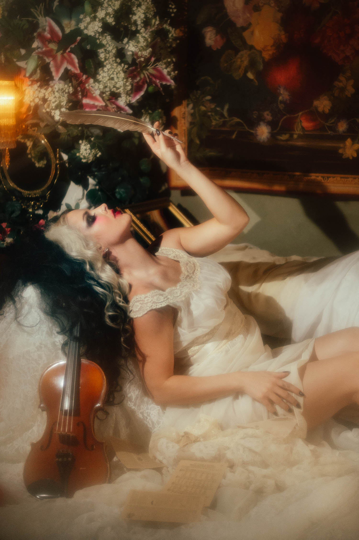 A model in an ivory lace nightgown reclines with a quill pen raised above her, a violin resting beside her amid soft florals in this dark academia photoshoot by Royal Lune Photo.