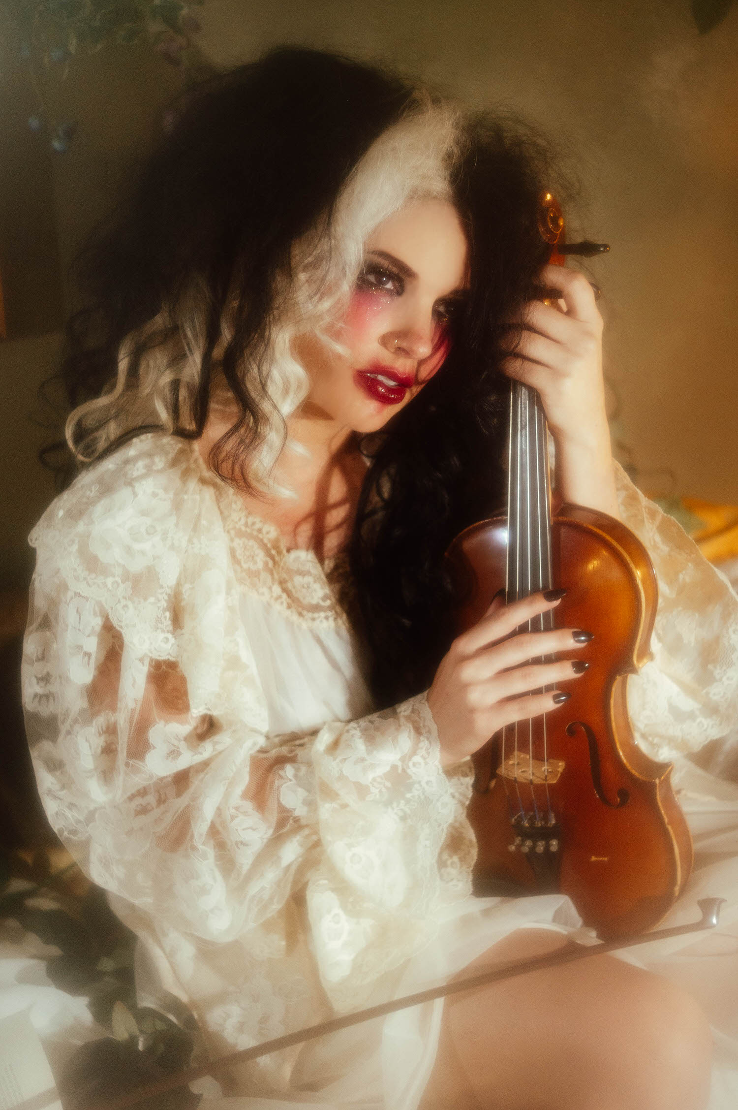 A model in an ivory lace robe holds a violin close to her face with dramatic red lipstick and glittery eye makeup in this dark academia photoshoot by Royal Lune Photo.