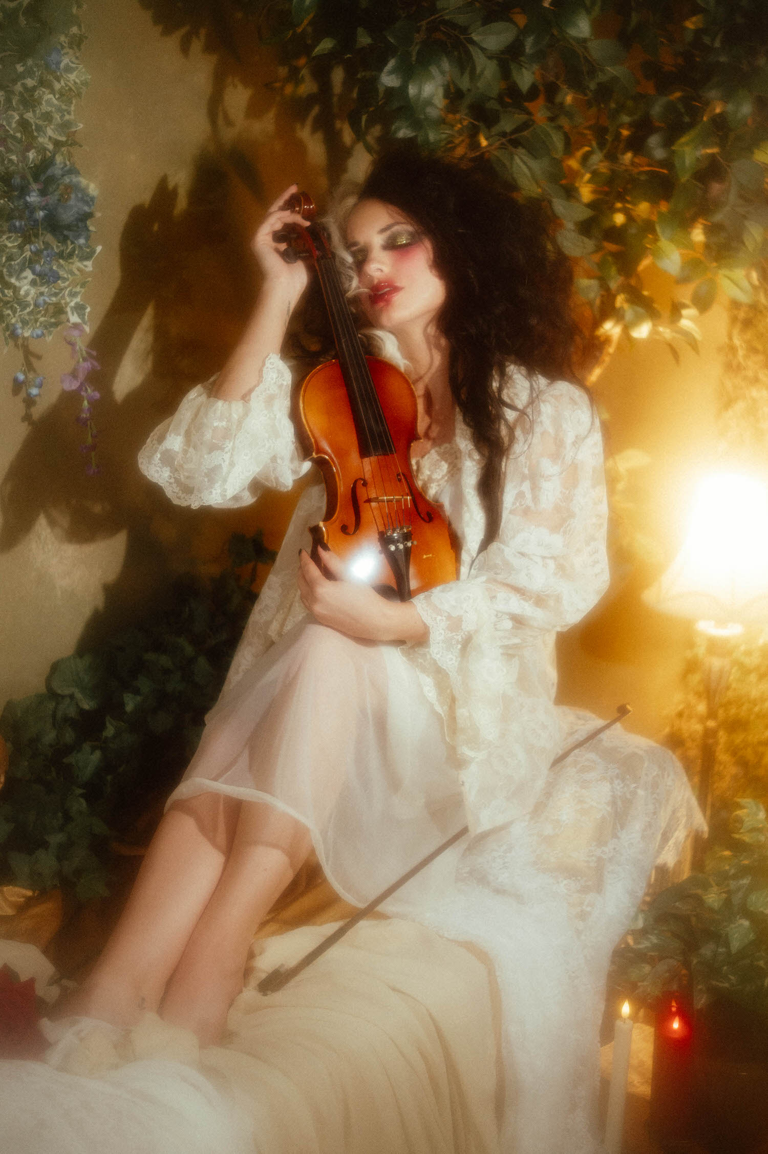 A model sits among greenery in a vintage ivory lace robe, cradling a violin and bow in this gothic boudoir photography session by Royal Lune Photo.