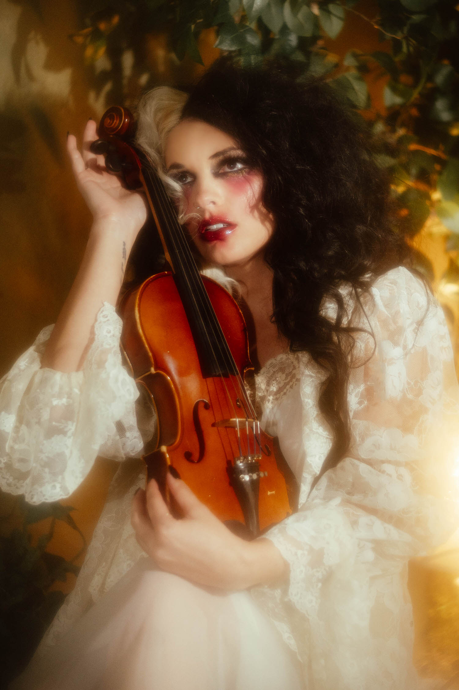 A close up portrait shows a model in an ivory lace robe holding a violin near her chin, framed by soft greenery and warm light in this fine art boudoir Dallas session by Royal Lune Photo.