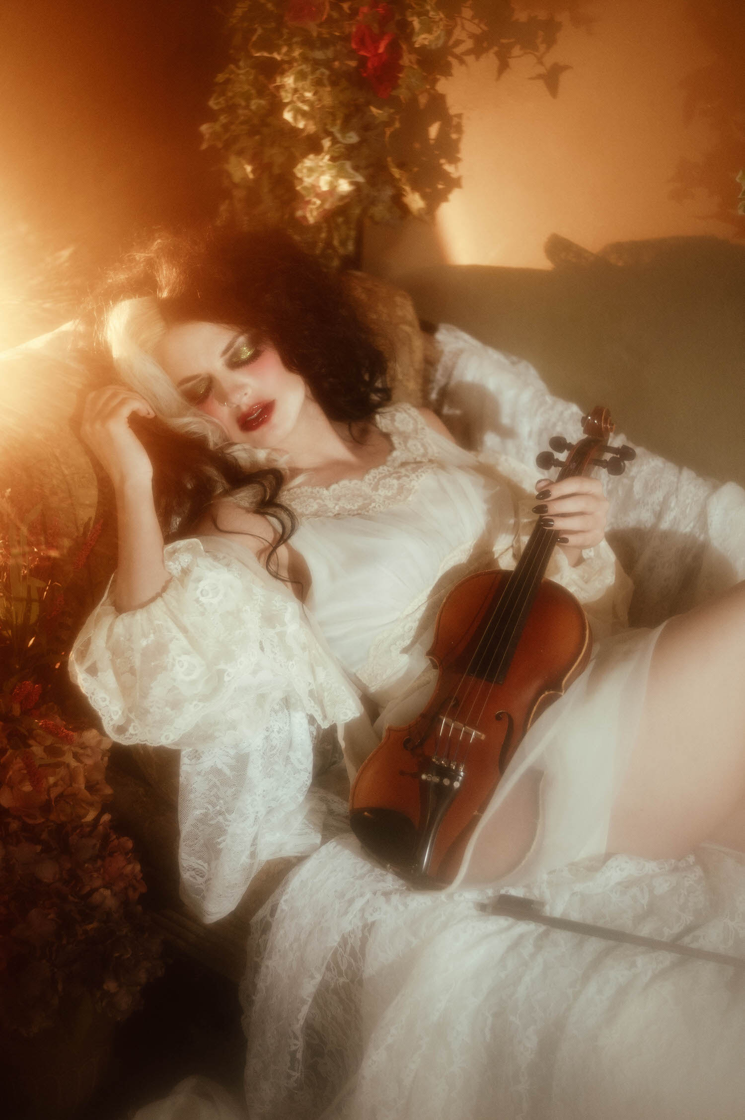 A model reclines on a lace covered daybed holding a violin, bathed in warm golden light with red flowers in the background in this dark academia photoshoot by Royal Lune Photo.