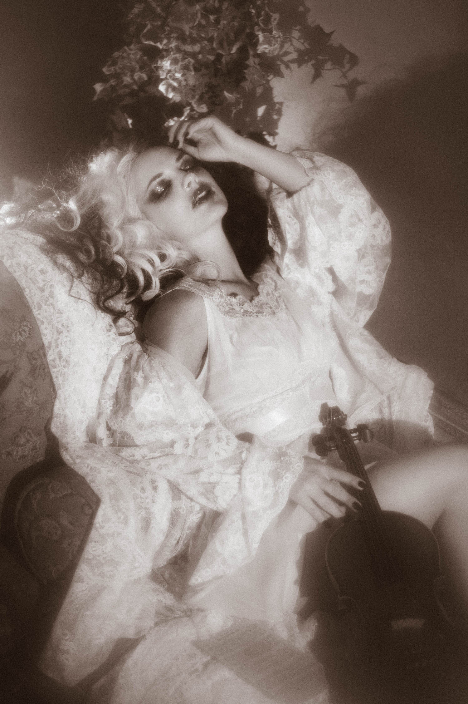A black and white portrait shows a model reclining with her arm raised to her hair while holding a violin, framed by ivy leaves in this moody poetry photoshoot by Royal Lune Photo.
