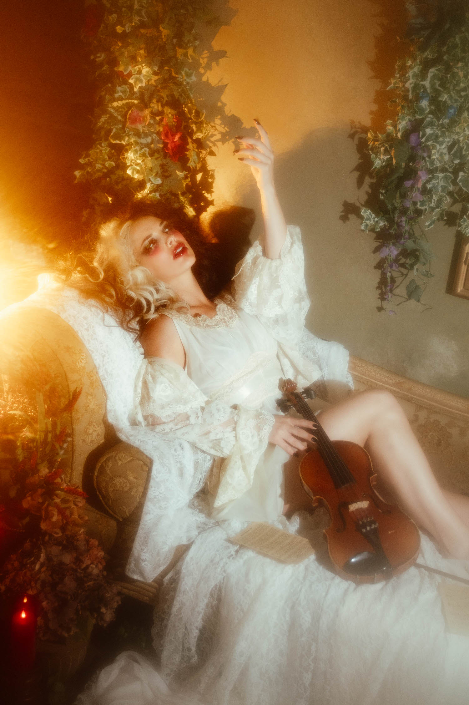 A model in a vintage ivory nightgown and lace robe sits with one arm reaching upward while holding a violin, surrounded by trailing greenery in this gothic boudoir photography portrait by Royal Lune Photo.
