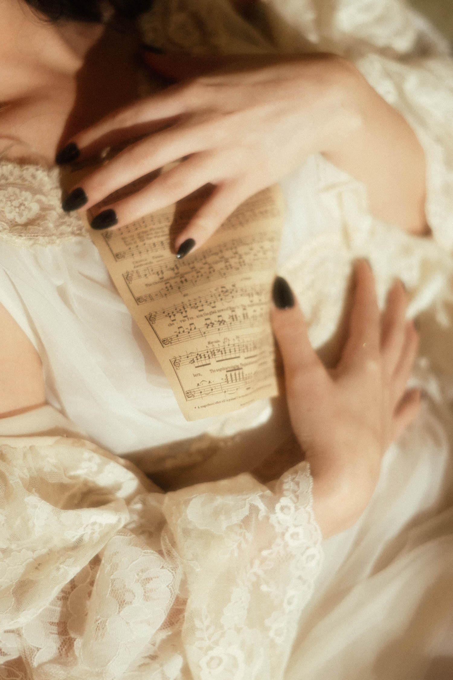 A close up image shows a model's hands with dark polished nails holding a page of antique sheet music against ivory lace fabric in this dark academia photoshoot by Royal Lune Photo.