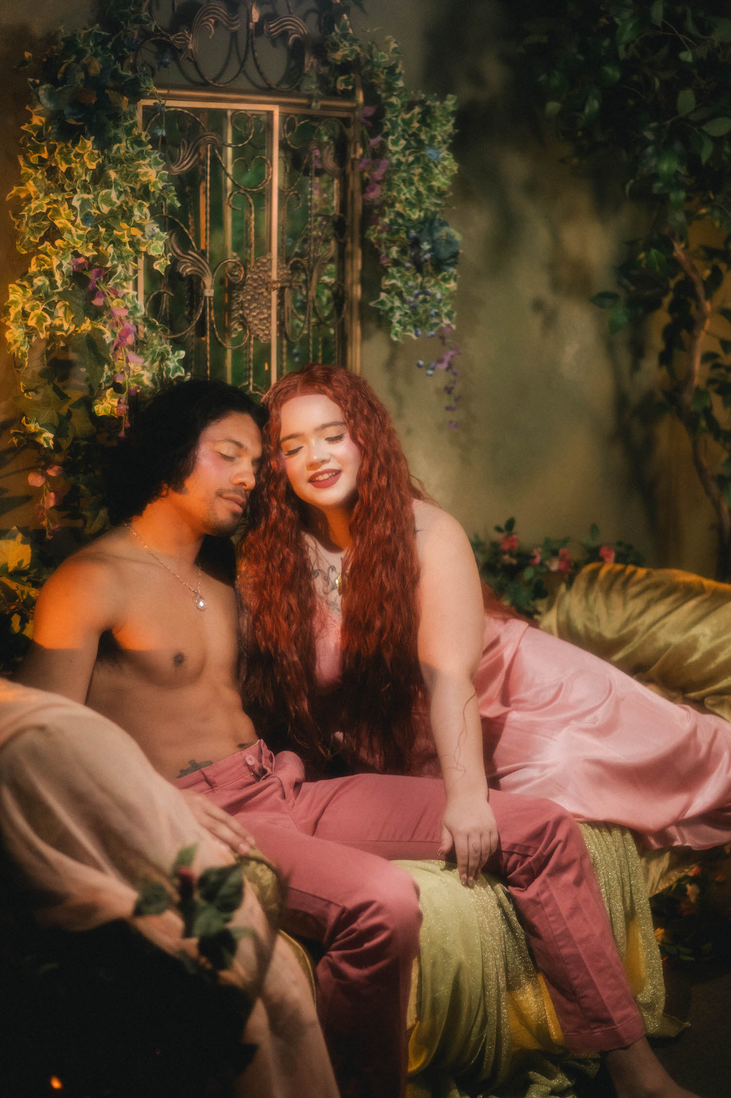 Dallas Fort Worth boudoir photographer captures a couple seated together on a gold and rose toned settee, surrounded by trailing ivy and lavender blooms against a wrought iron backdrop. Royal Lune Photo