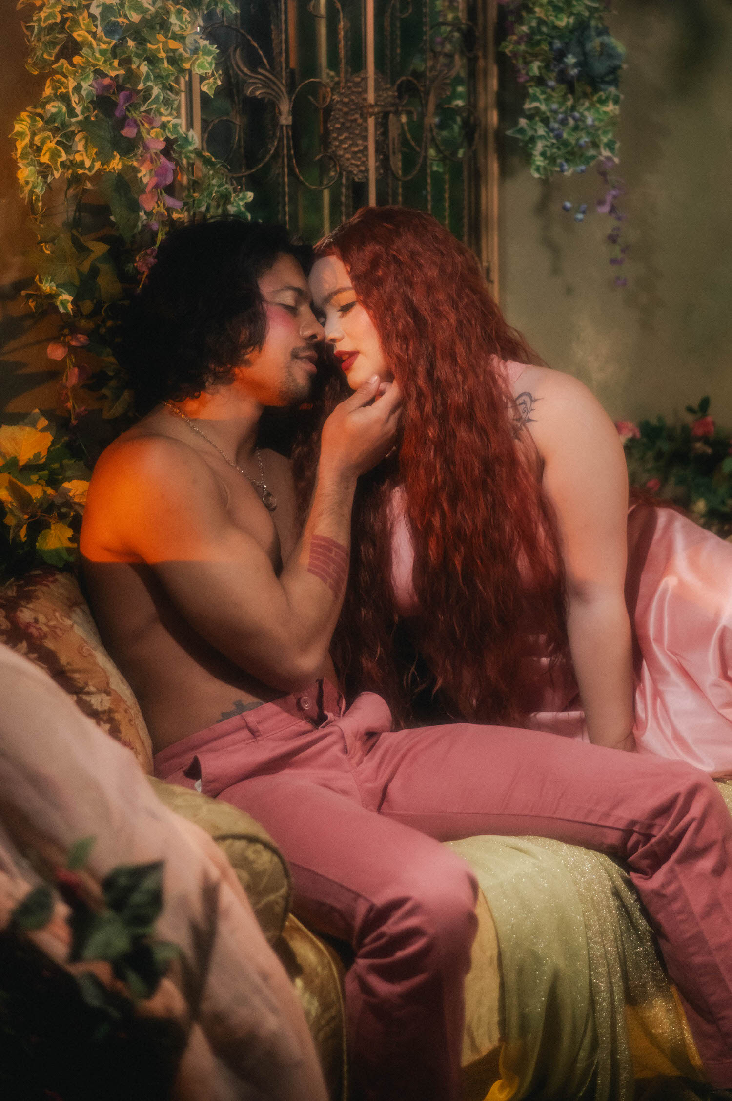 A queer couples boudoir portrait shows two figures leaning into a tender moment on a floral draped bed, warm ambient light catching the folds of a satin robe. Royal Lune Photo