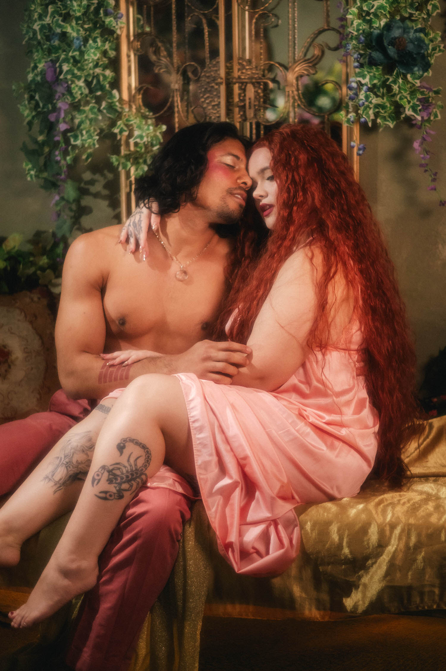 Couples boudoir photoshoot ideas come to life as a woman in a flowing coral silk gown sits across her partner's lap, a scorpion tattoo visible along his thigh amid gold brocade pillows. Royal Lune Photo