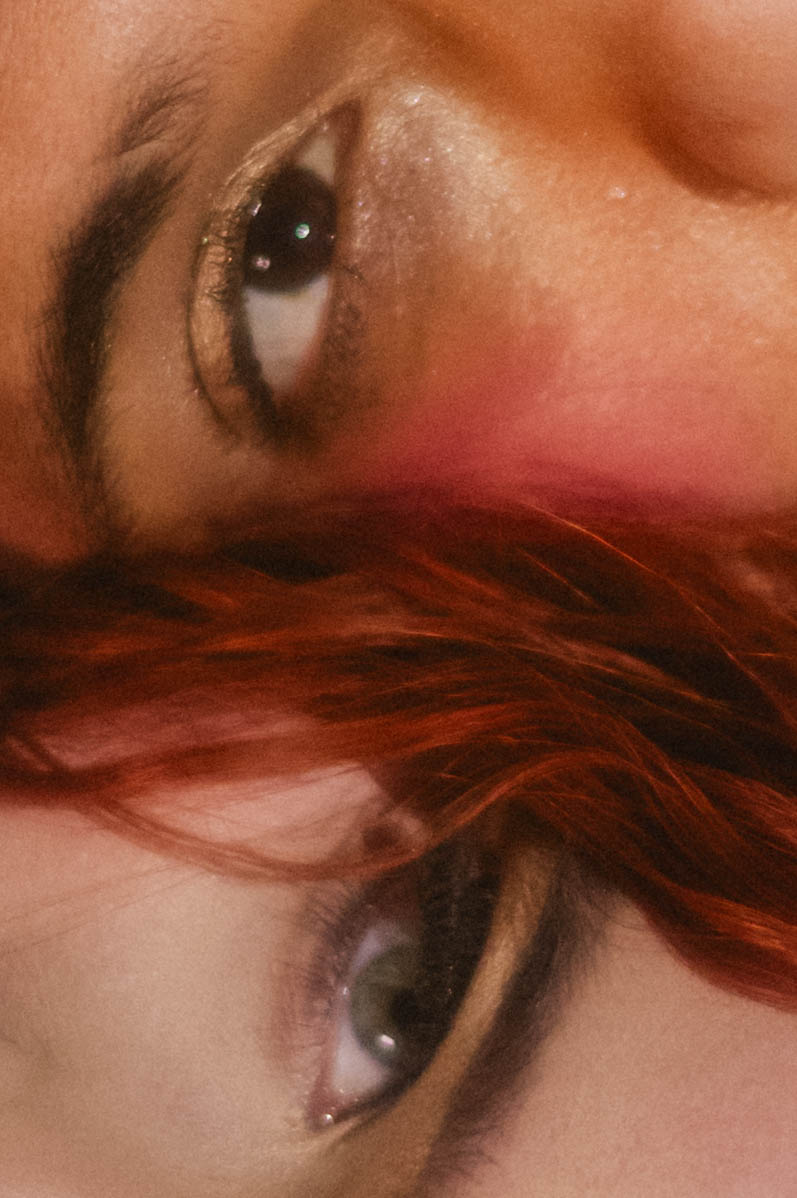Extreme close up detail shot from a Renaissance boudoir photoshoot capturing two eyes meeting through a veil of red hair, warm blush tones across both faces. Royal Lune Photo