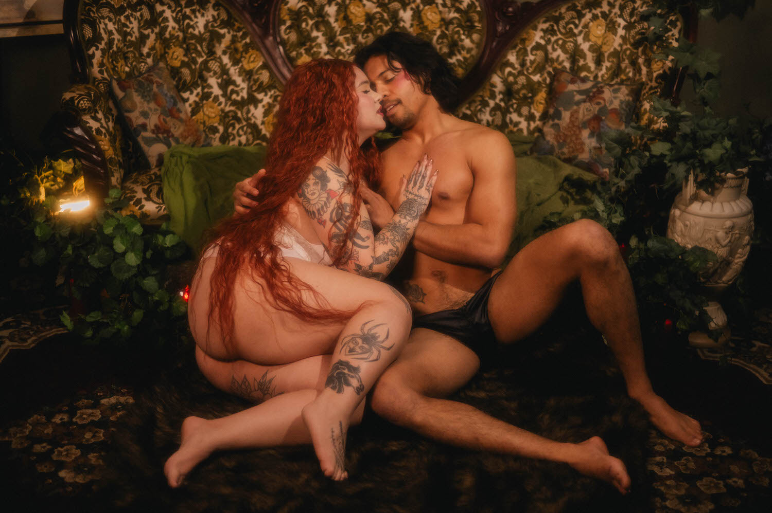 Wide shot from a couples boudoir photoshoot inside a vintage parlor setting, the pair seated together on a floral antique sofa beside a classical stone urn. Royal Lune Photo