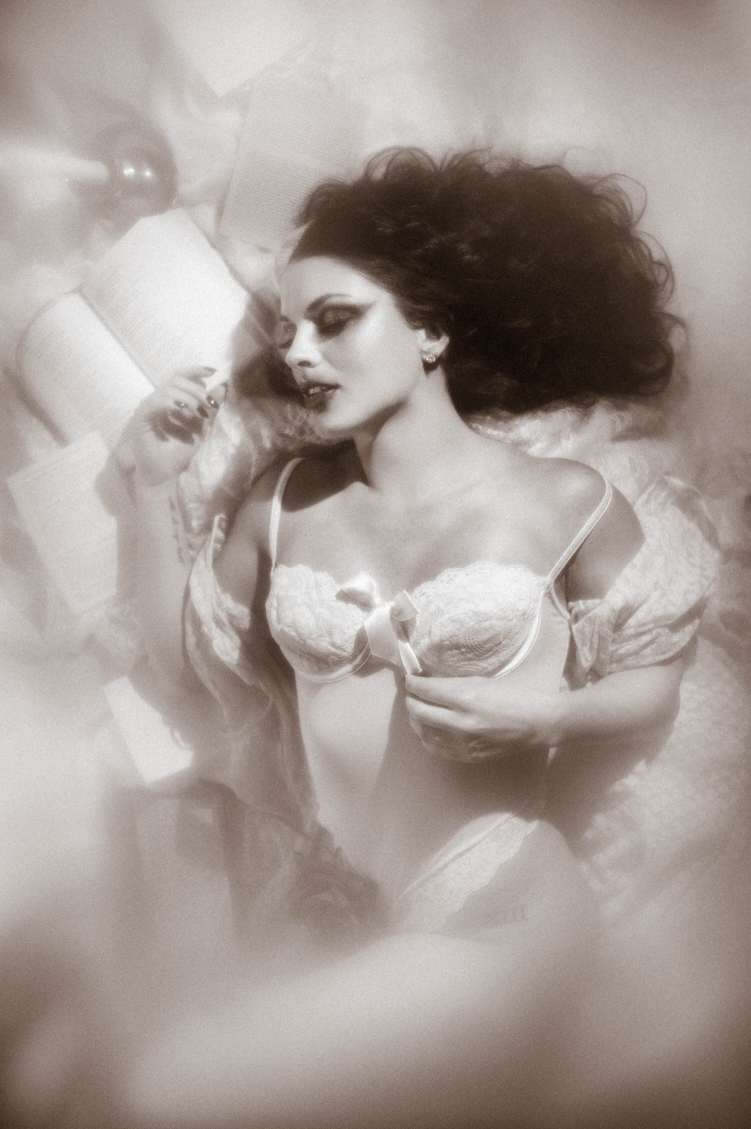 A black and white portrait shows a dark haired model in a vintage ivory lace bodysuit reaching toward an open book beside a lit candle in this moody poetry photoshoot at Royal Lune Studio by Royal Lune Photo.