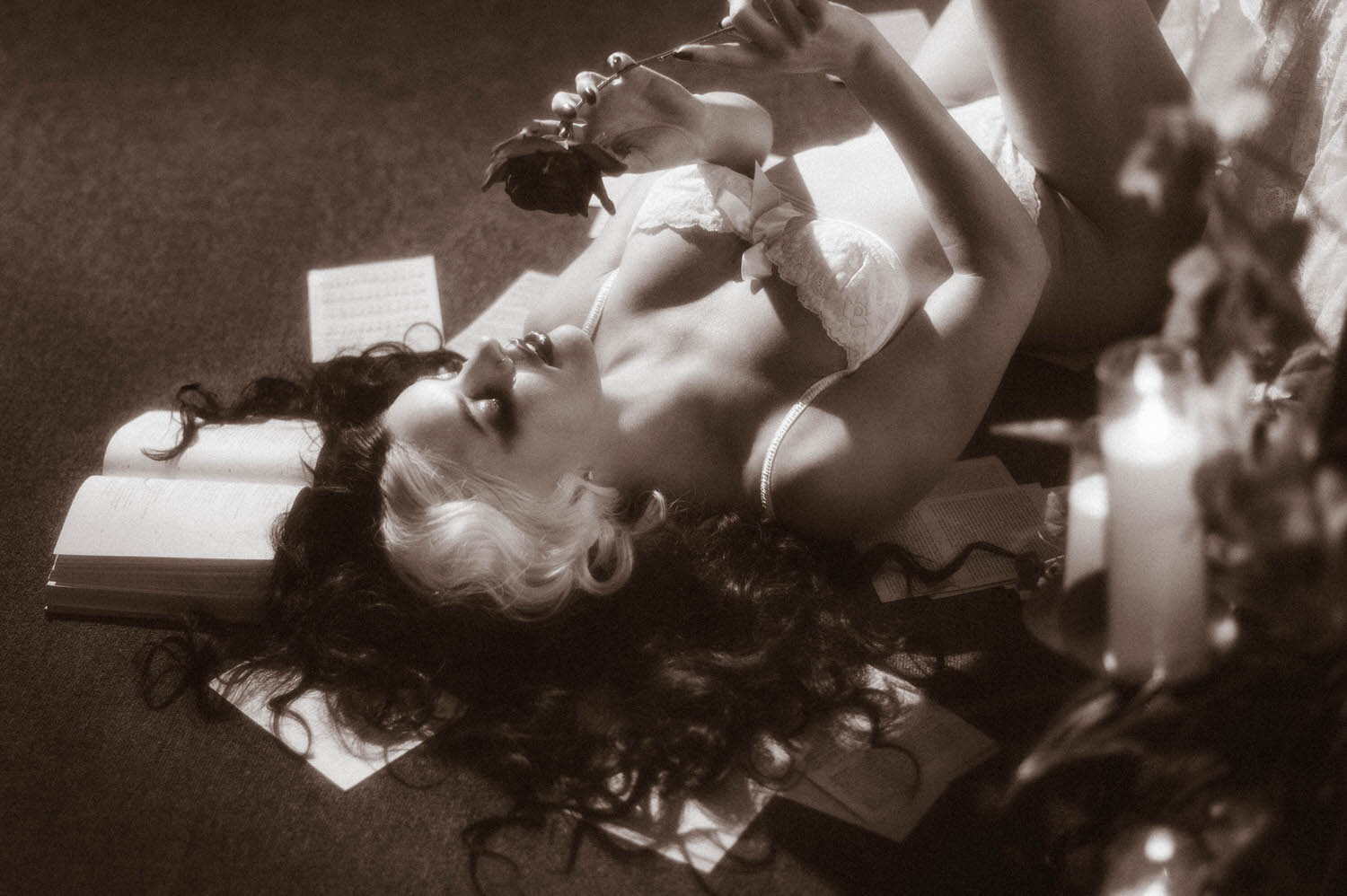 A black and white image shows a model reclining with a rose held above an open book and a lit candle in this moody poetry photoshoot by Royal Lune Photo.