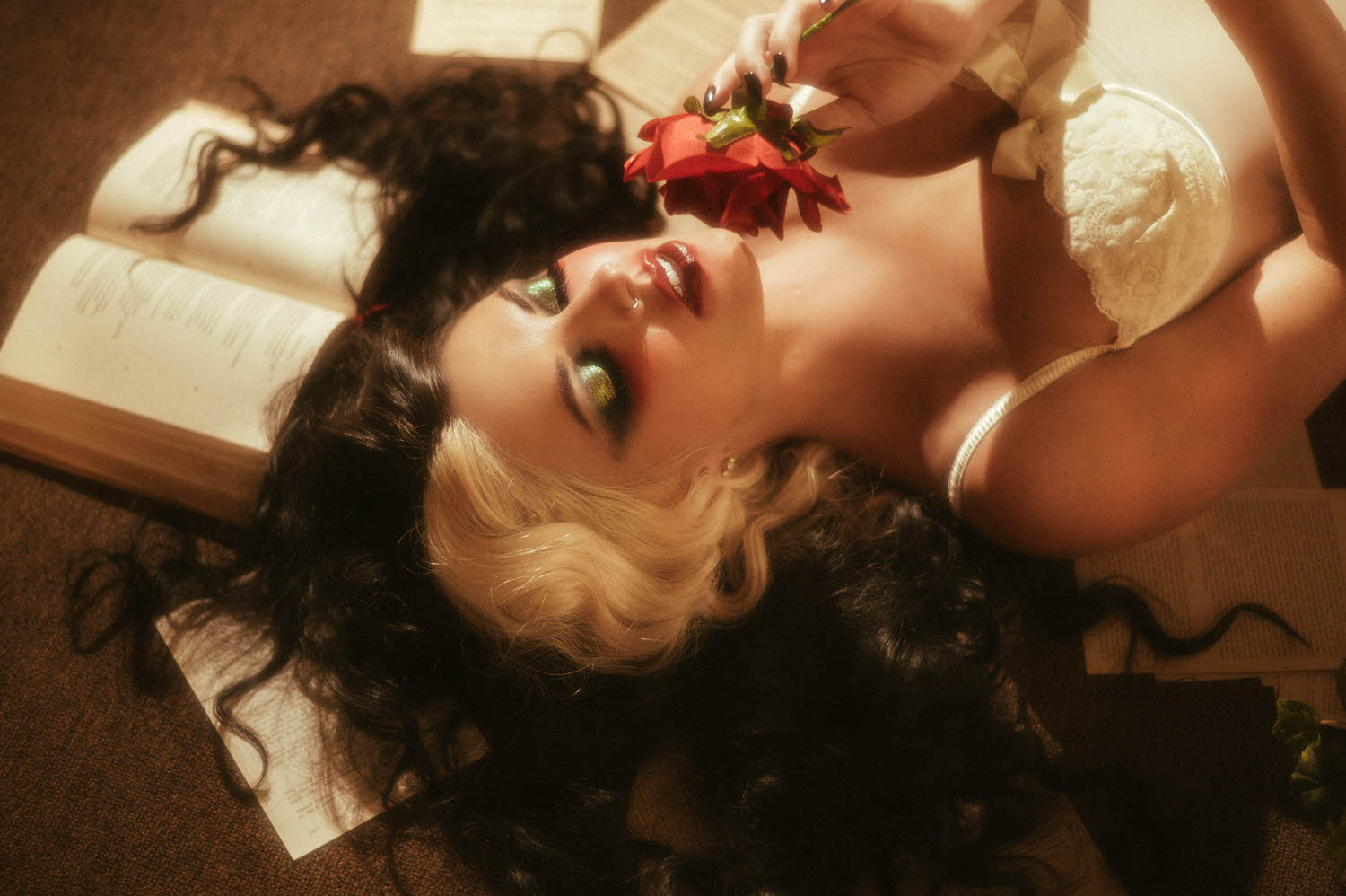 A close up portrait captures a model with shimmering green eyeshadow and red lips holding a red rose above an open book in this heartbreak photoshoot idea by Royal Lune Photo.