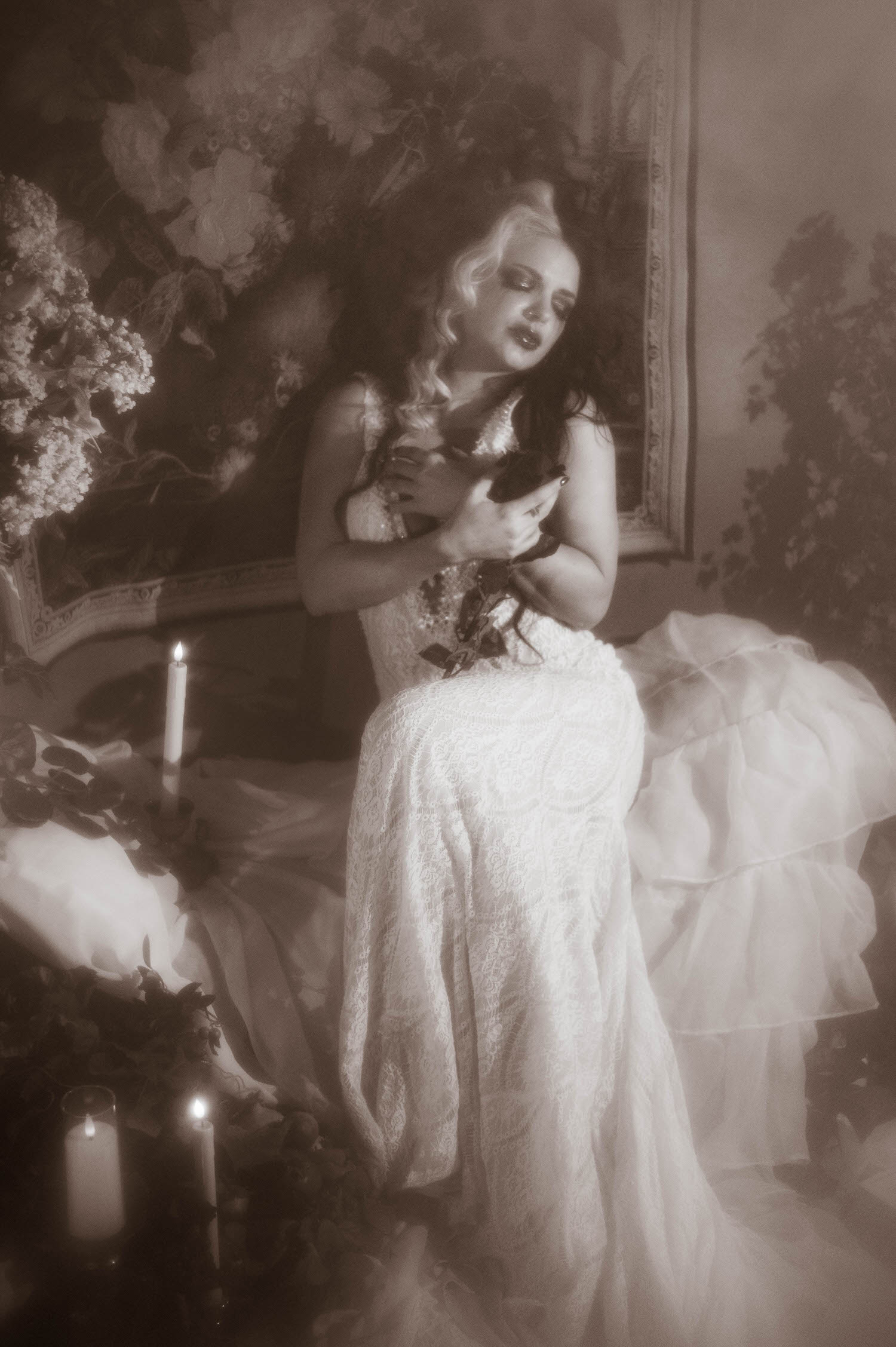 A black and white portrait shows a model in a lace wedding gown seated while holding a black rose close to her chest in this moody poetry photoshoot by Royal Lune Photo.