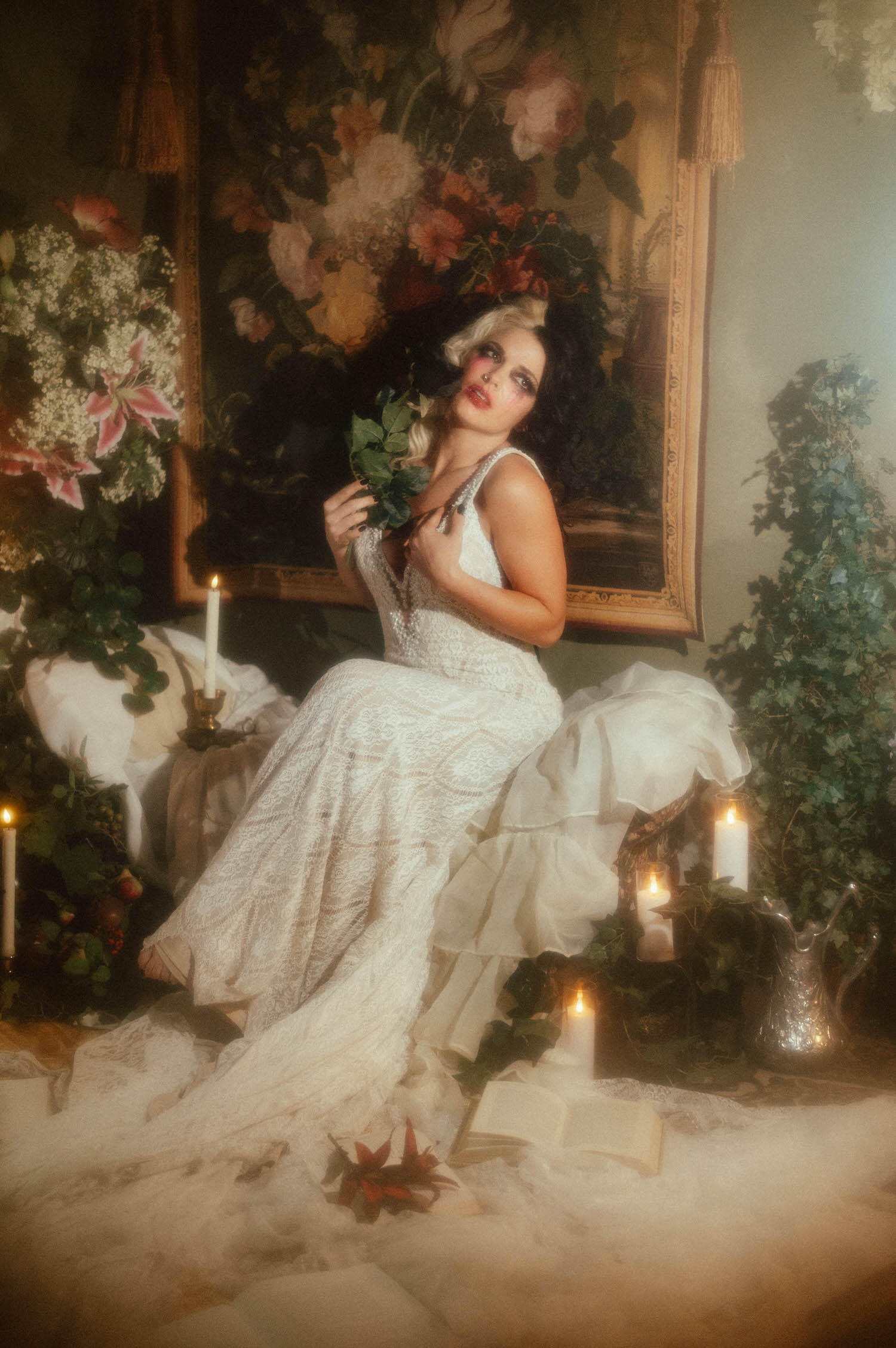 A model in a Madison James lace wedding gown sits before a floral tapestry holding greenery, surrounded by candlelight in this Tortured Poets Department photoshoot by Royal Lune Photo.