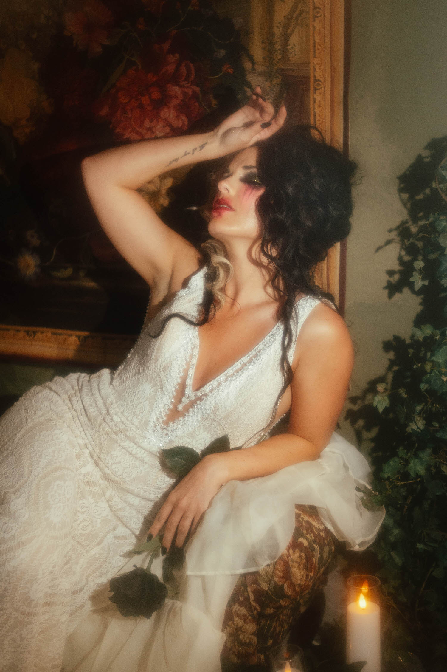 A model in a Madison James wedding gown sits with one arm raised while holding a black rose in her lap in this dark academia photoshoot by Royal Lune Photo.