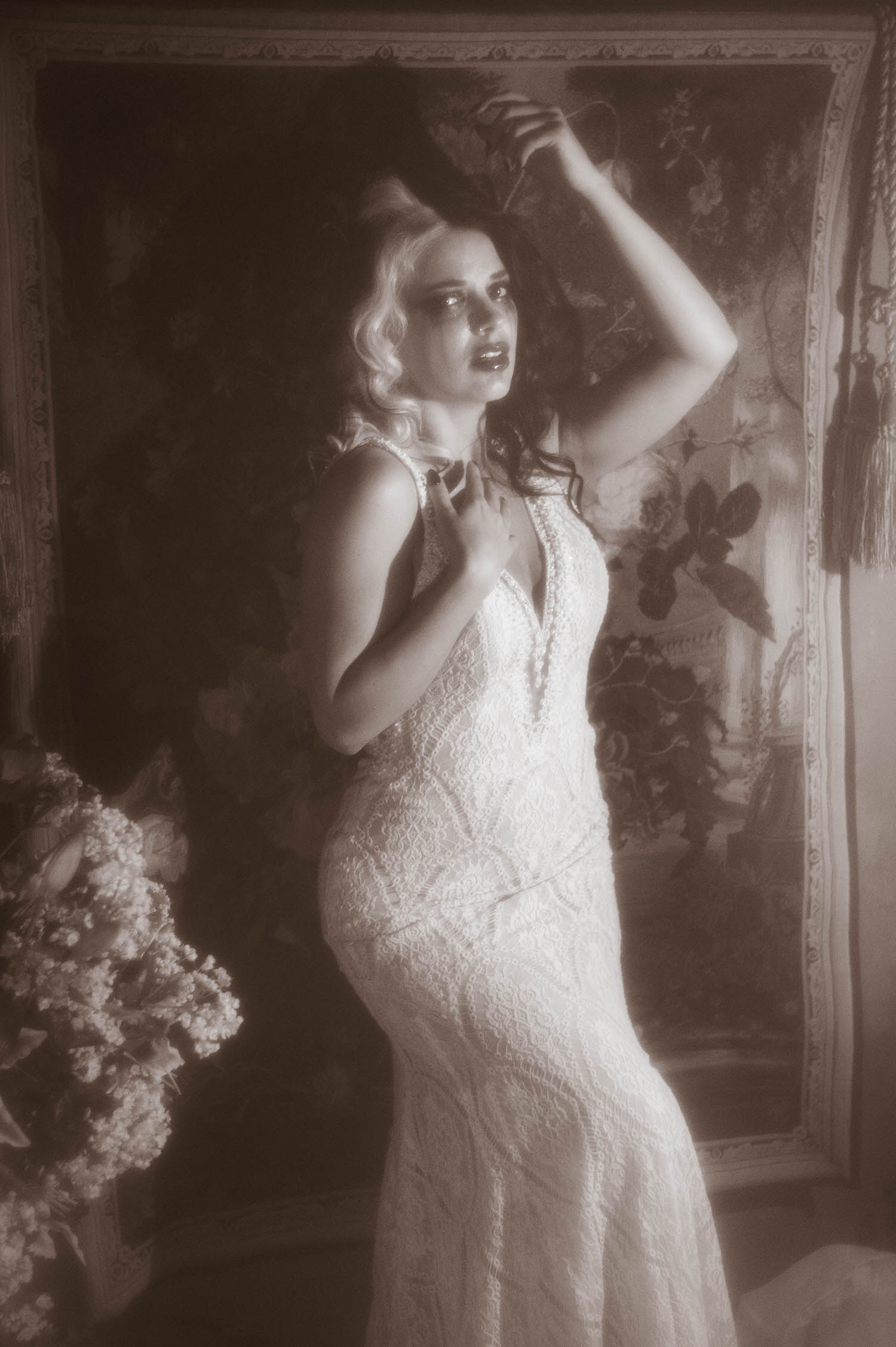 A black and white portrait shows a model in a fitted lace wedding gown standing with one hand raised near her hair in this fine art boudoir Dallas session by Royal Lune Photo.