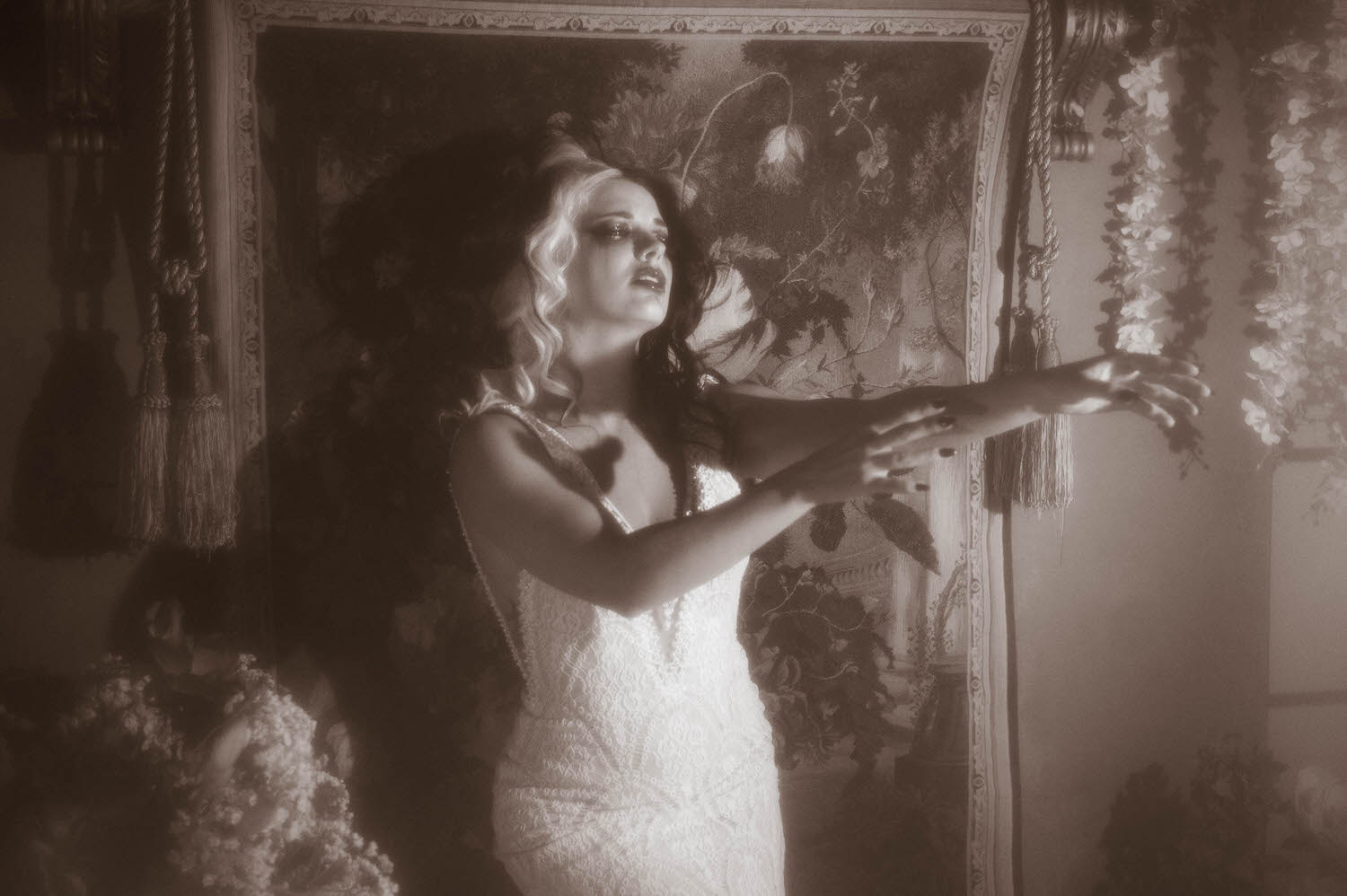 A black and white portrait shows a model in a lace wedding gown standing with both arms outstretched toward hanging tassels and flowers in this Tortured Poets Department photoshoot by Royal Lune Photo.