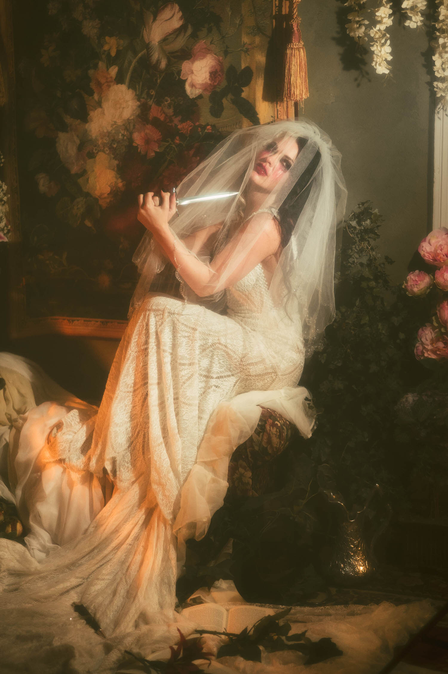 A model in a lace wedding gown holds a dagger beneath a sheer veil, framed by pink flowers and candlelight in this dark academia photoshoot by Royal Lune Photo.