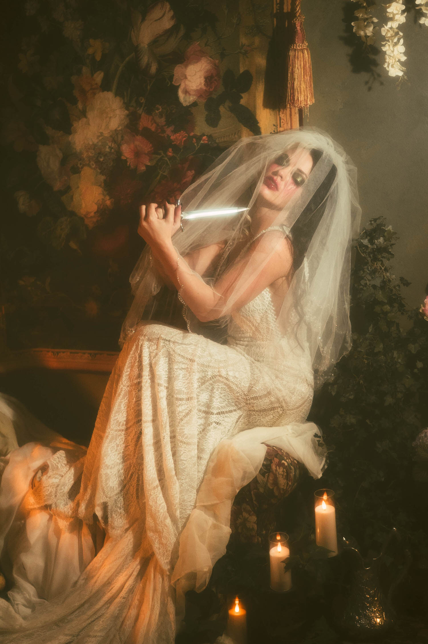 A model in a lace wedding gown and sheer veil sits holding a dagger near her face, surrounded by candlelight and floral tapestry in this Tortured Poets Department photoshoot by Royal Lune Photo.