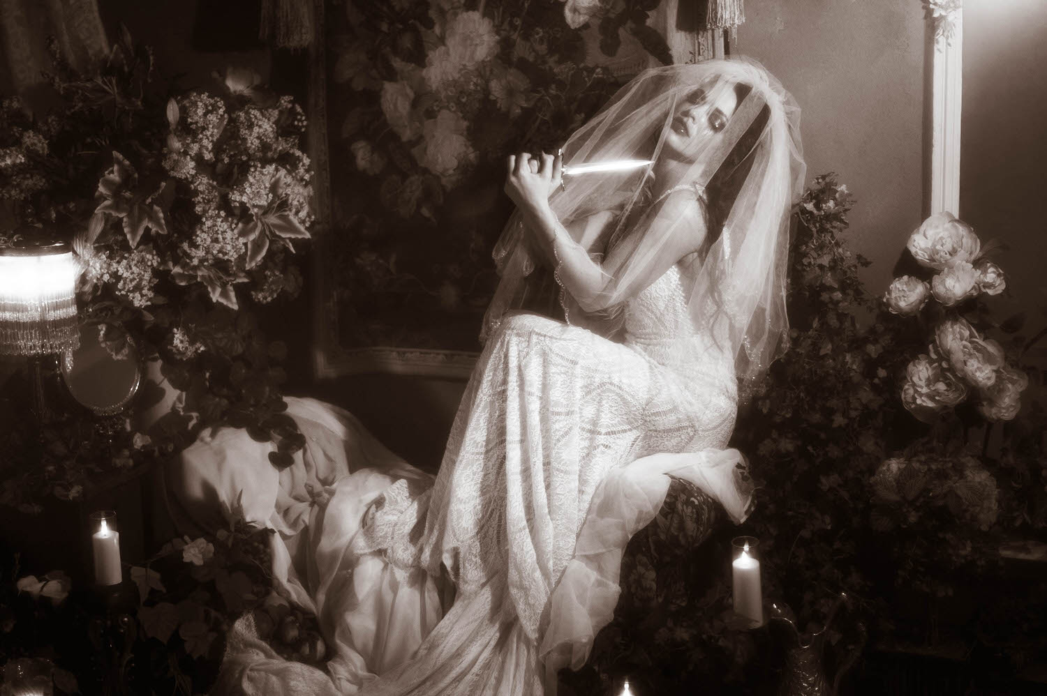 A black and white portrait shows a model in a lace wedding gown holding a dagger beneath a sheer veil against a floral tapestry backdrop in this gothic boudoir photography session by Royal Lune Photo.