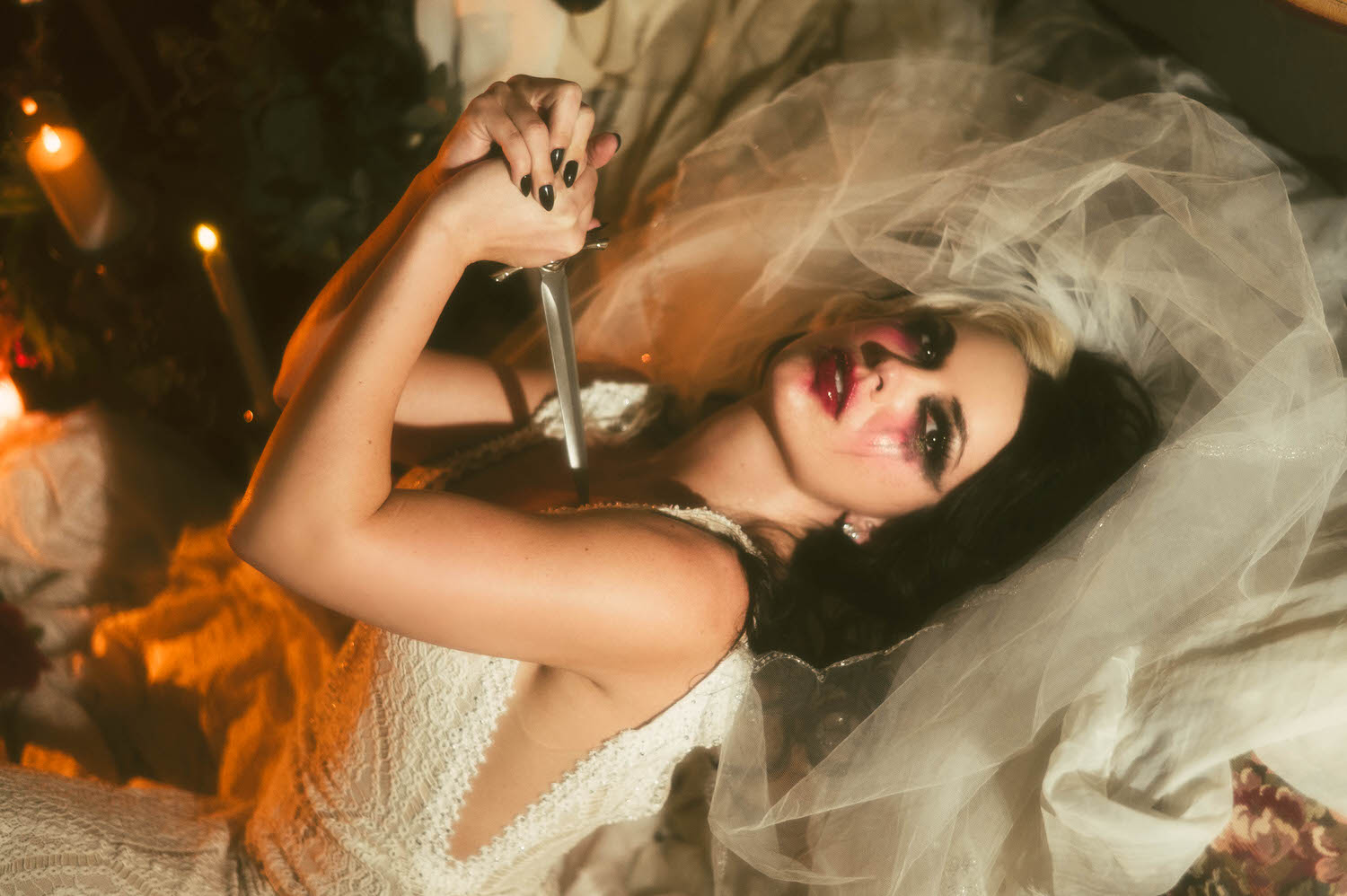 A close up portrait shows a model in a lace wedding gown holding a dagger to her chest beneath a sheer veil in this Tortured Poets Department photoshoot by Royal Lune Photo.