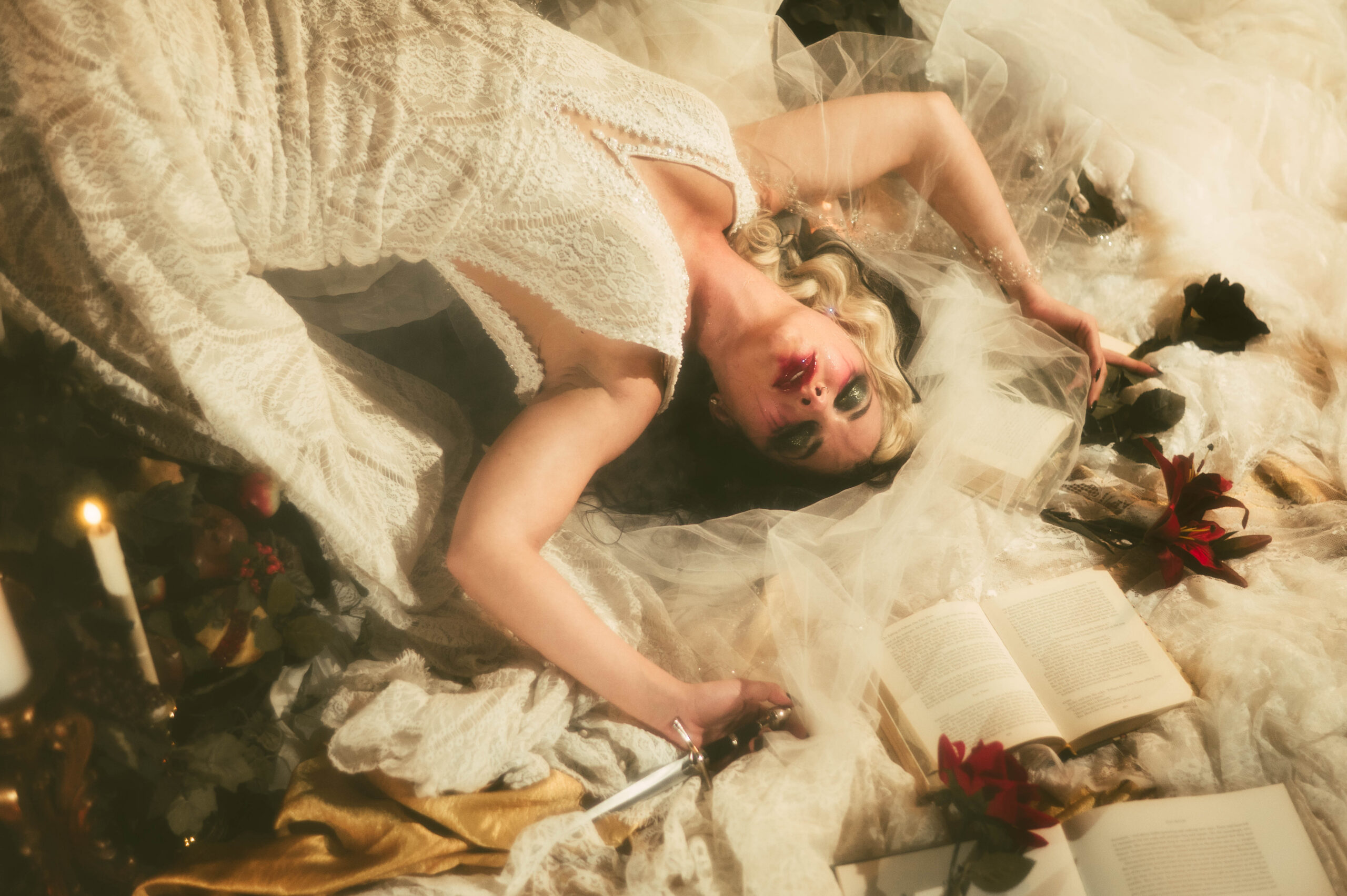 Woman in a white lace dress lies on the floor with blood on her mouth, surrounded by books, candles, flowers, and a dagger—a hauntingly beautiful dark academia photoshoot inspired by Taylor Swift album themes.