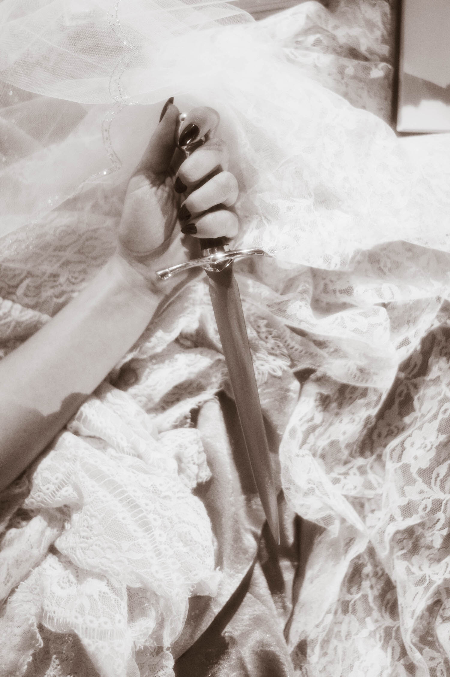 A black and white close up image shows a model's hand with dark polished nails gripping a dagger against lace fabric and a sheer veil in this heartbreak photoshoot idea by Royal Lune Photo.