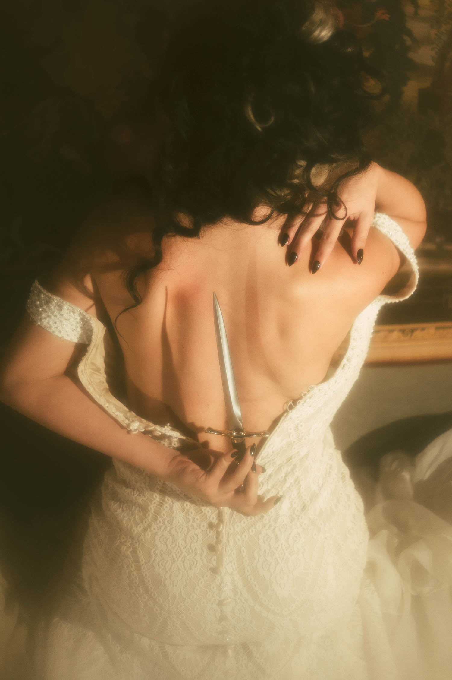 A close up image shows a model's bare back and dark curled hair as she holds a dagger against her spine in this dark academia photoshoot by Royal Lune Photo.