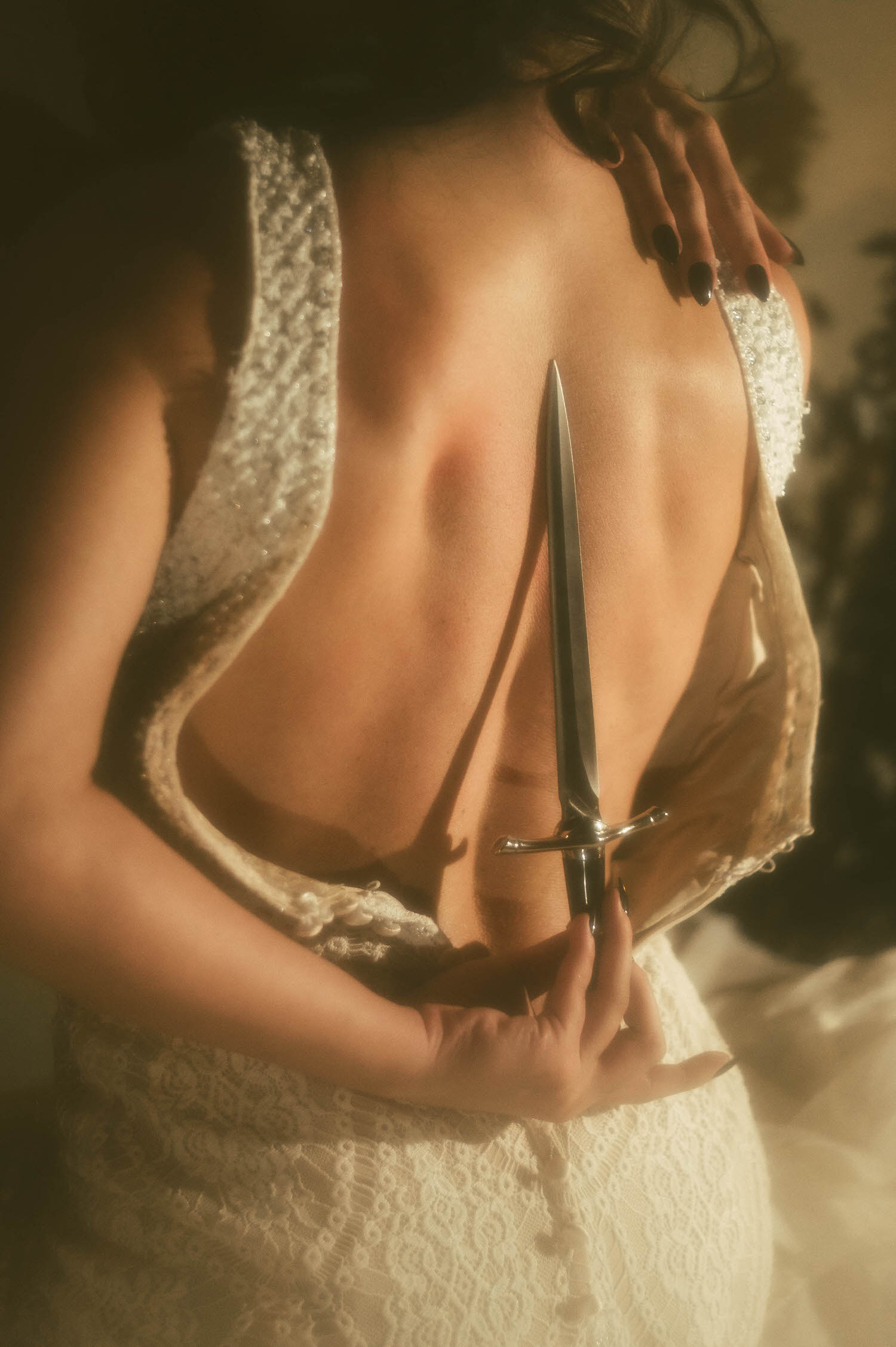 A close up image shows a model's bare back in a beaded lace wedding gown with a dagger held vertically against her spine in this Tortured Poets Department photoshoot by Royal Lune Photo.