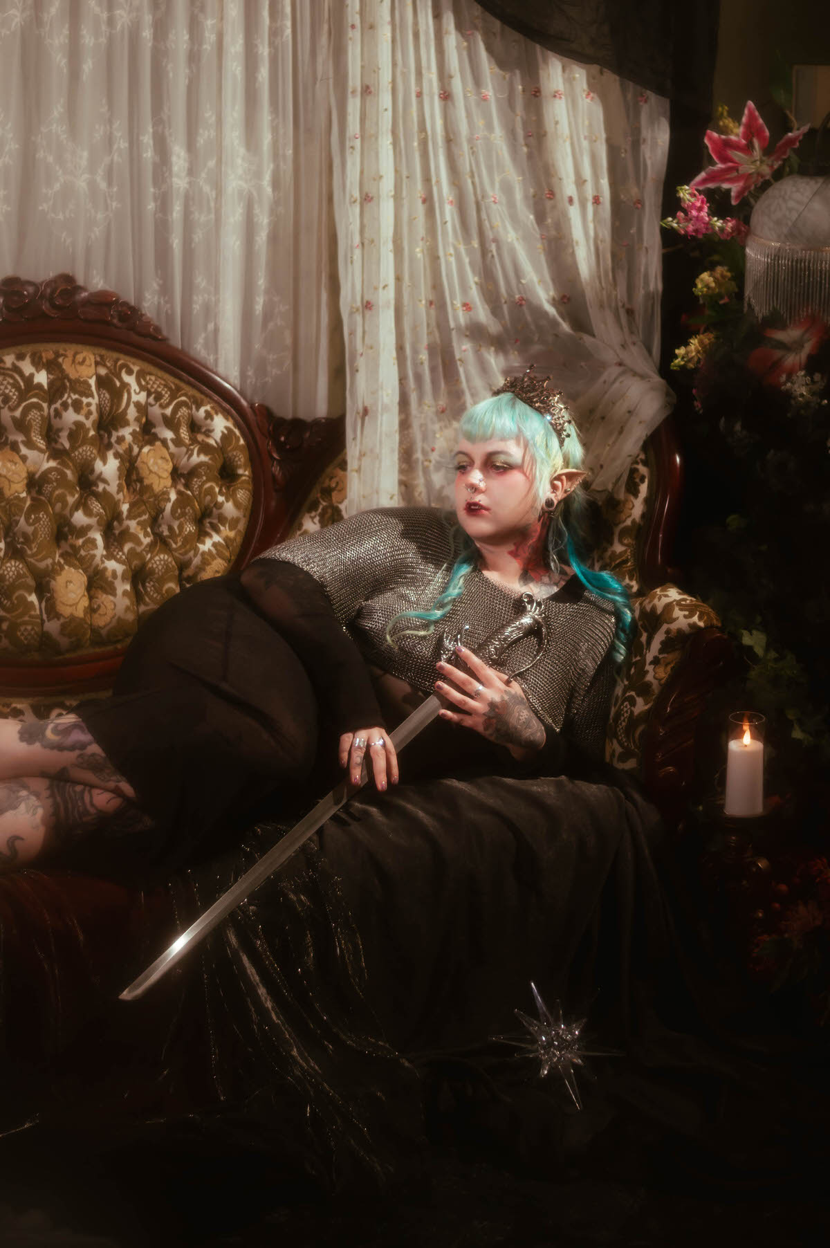 A Dallas Fort Worth branding photographer image of a woman reclining on a floral tufted sofa with eyes closed, wearing a dark tiara and chainmail top with a red patterned fabric draped beside her, Royal Lune Photo.