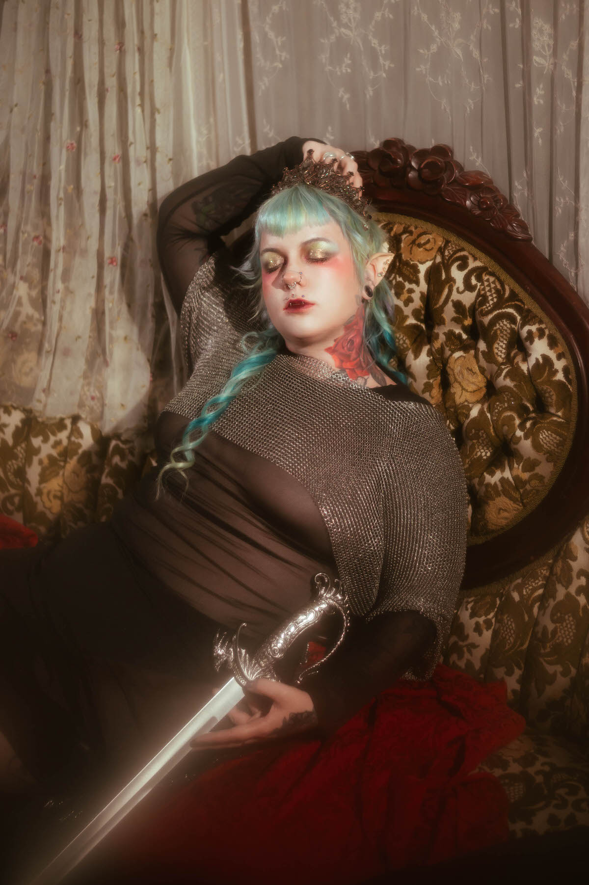 A black and white tattoo artist branding photoshoot portrait of a woman reclining on a tufted sofa holding a sword upright, wearing a chainmail top and jeweled tiara, Royal Lune Photo.