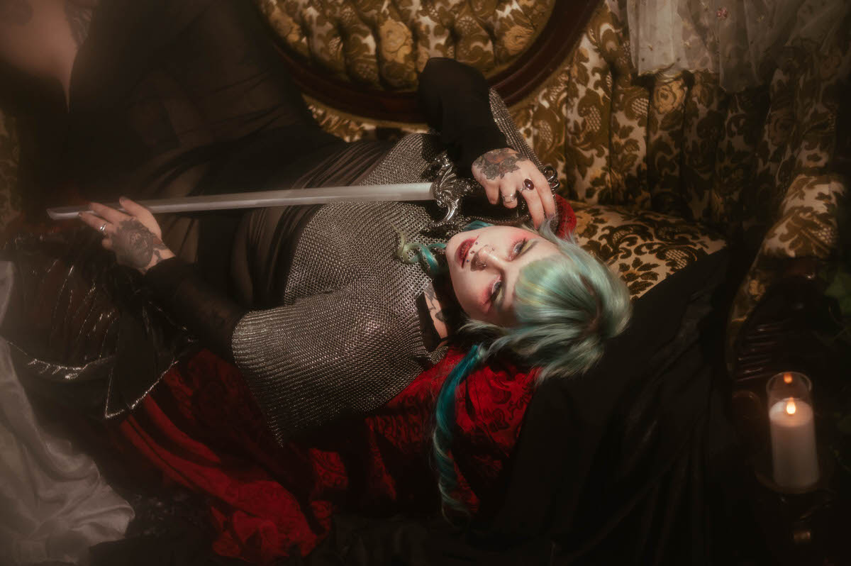 A close up tattoo artist branding photography image of a woman holding a sword hilt near her face while lying against red brocade fabric, wearing a chainmail top with visible piercings, Royal Lune Photo.