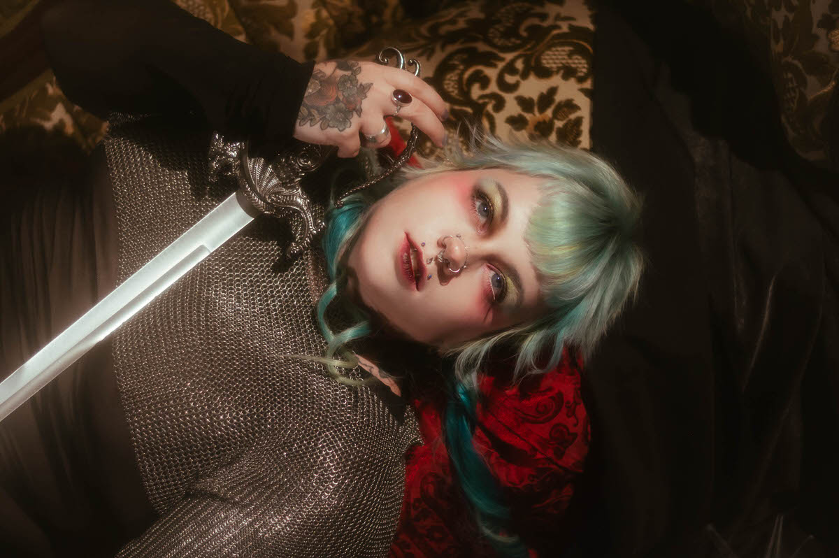 A content photography for creatives in Dallas close up image of a woman lying back with lips parted, holding a sword hilt near her shoulder, gold and pink eye makeup visible against red fabric, Royal Lune Photo.