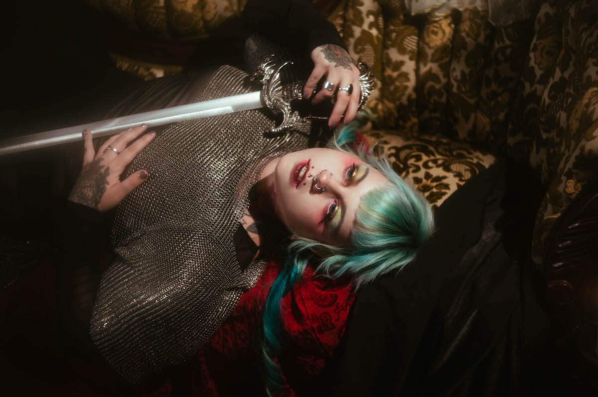 A tattoo artist branding photoshoot close up image of a woman lying back with a glowing bladed sword held near her chainmail top, red patterned fabric visible beneath her, Royal Lune Photo.