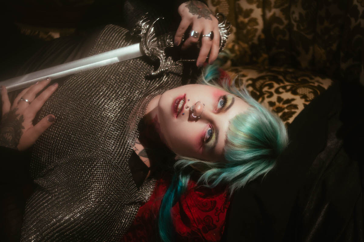 A personal branding photos for artists close up portrait of a woman with teal hair in twin braids, holding a sword hilt near her cheek while wearing a metallic chainmail top, Royal Lune Photo.