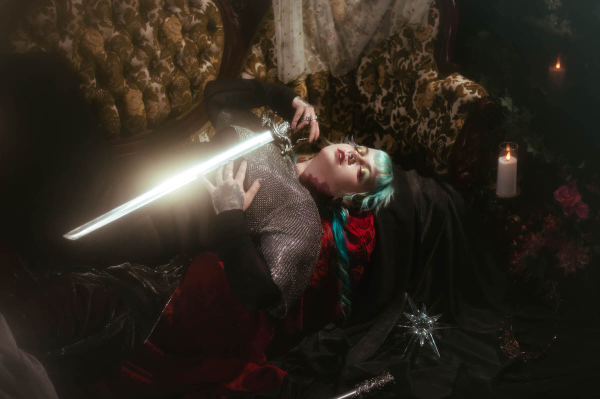 A tattoo artist branding photoshoot close up image of a woman lying back with a glowing bladed sword held near her chainmail top, red patterned fabric visible beneath her, Royal Lune Photo.